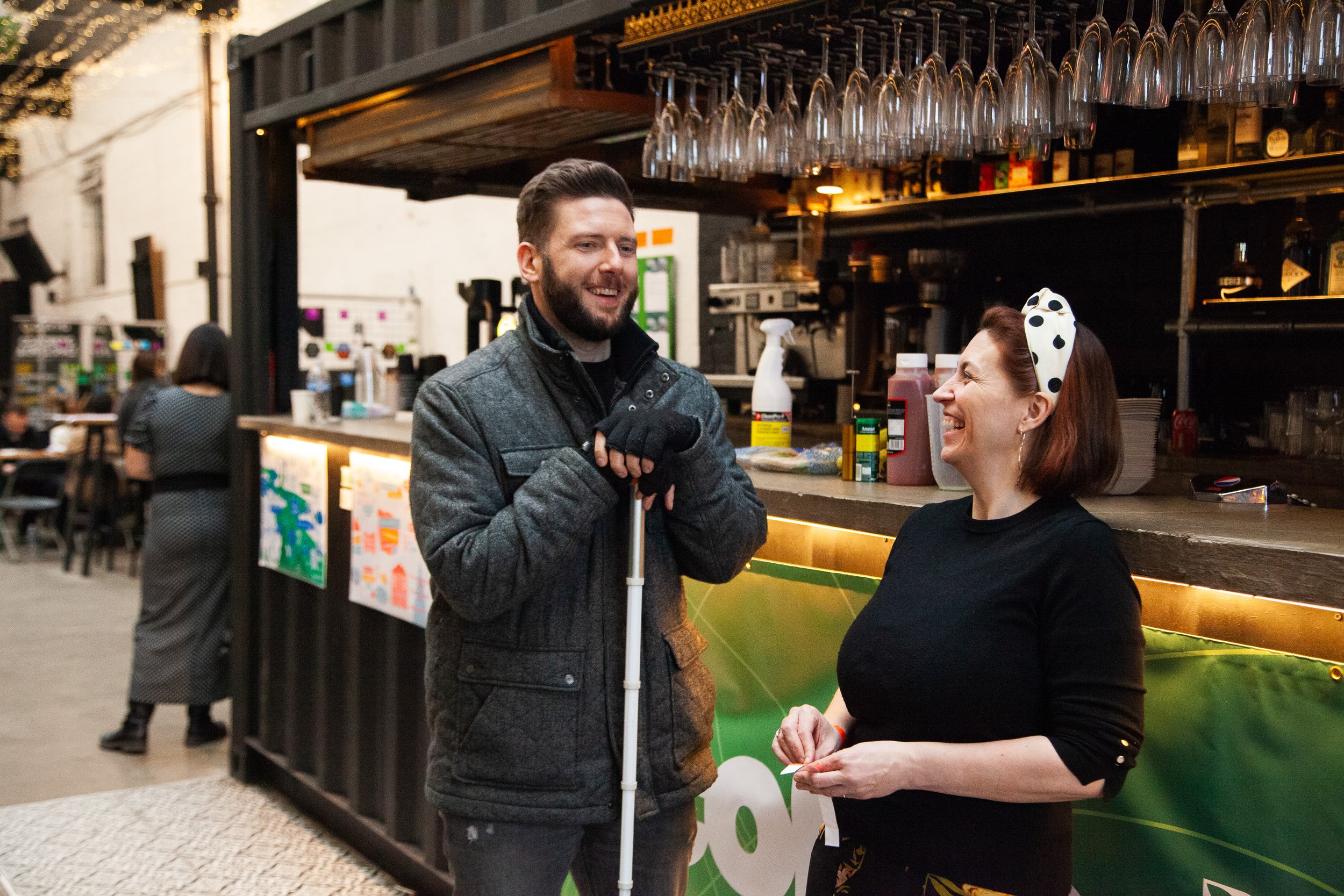A man with a cane and a woman with a polka dot headband sharing a laugh at an outdoor bar or café.