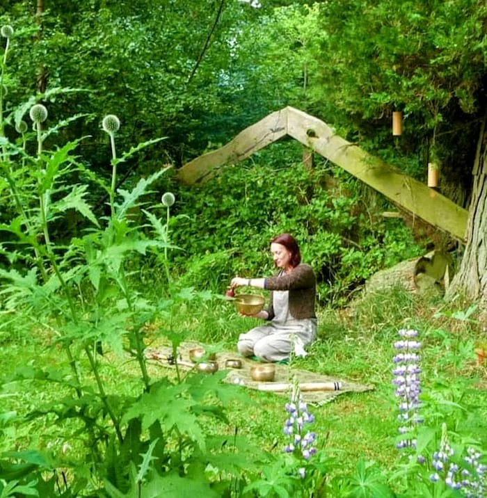 A woman in outdoor clothing kneeling on a mat in a lush green garden, holding a bowl and utensil, surrounded by various plants, trees, and flowers, with a wooden structure and trees in the background.
