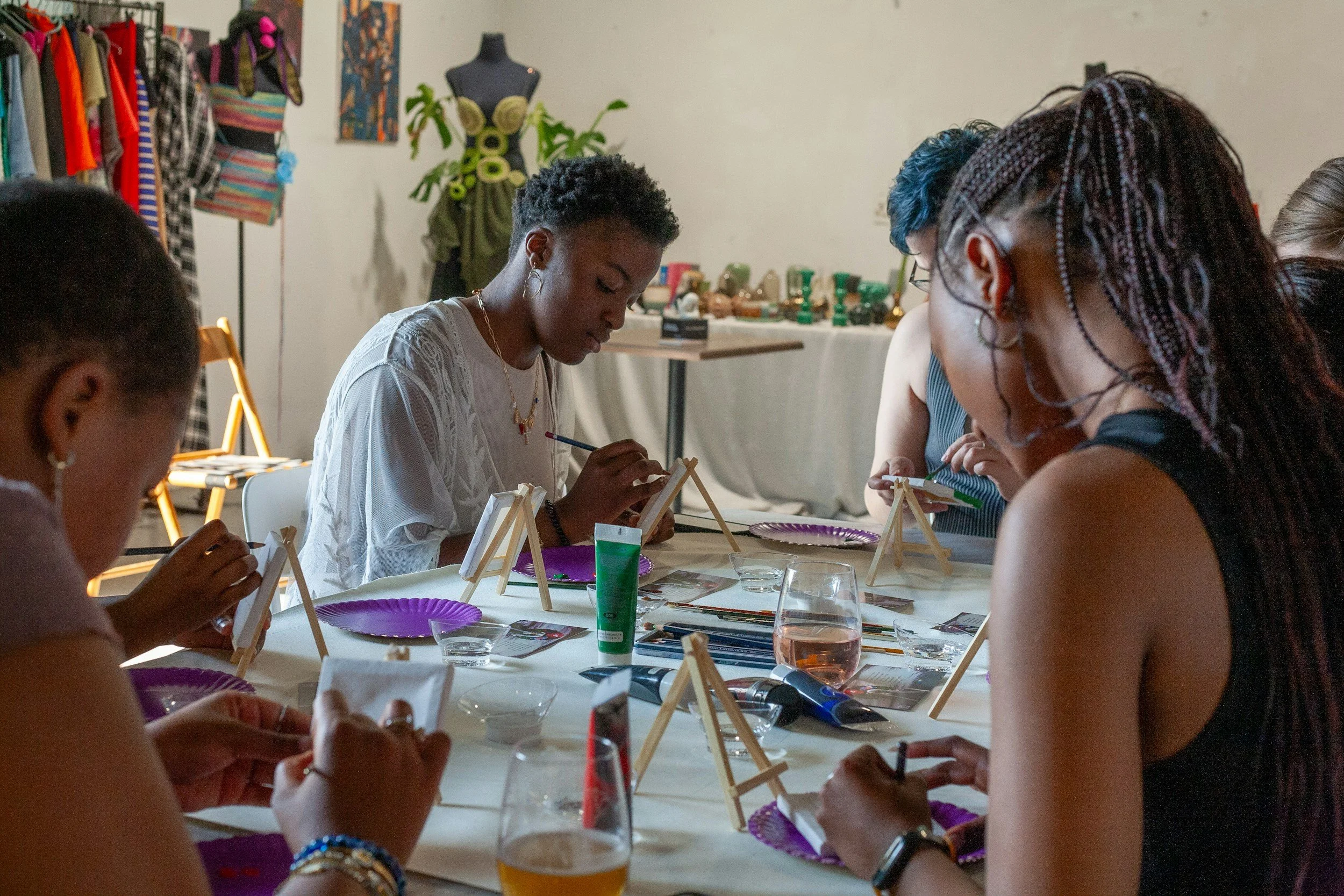 Group of women sitting around a table painting mini canvases on small easels for an art class, with art supplies on the table, in a room decorated with clothing and craft items.