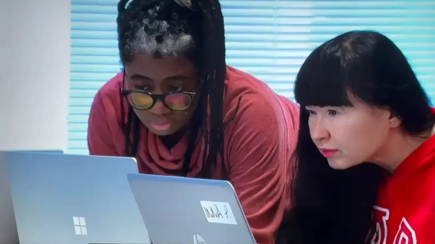 Two women looking at a laptop computer. One woman wears glasses and a red shirt, the other has long dark hair and is dressed in red. They are focused on the screen in a room with window blinds.