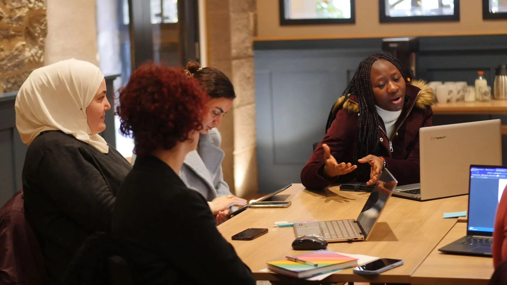 Four women sitting at a wooden table in a meeting room, engaged in discussion. One woman is speaking with expressive hand gestures. The table has laptops, a smartphone, a mouse, notebooks, and pens. The room has windows and a brick wall in the background.