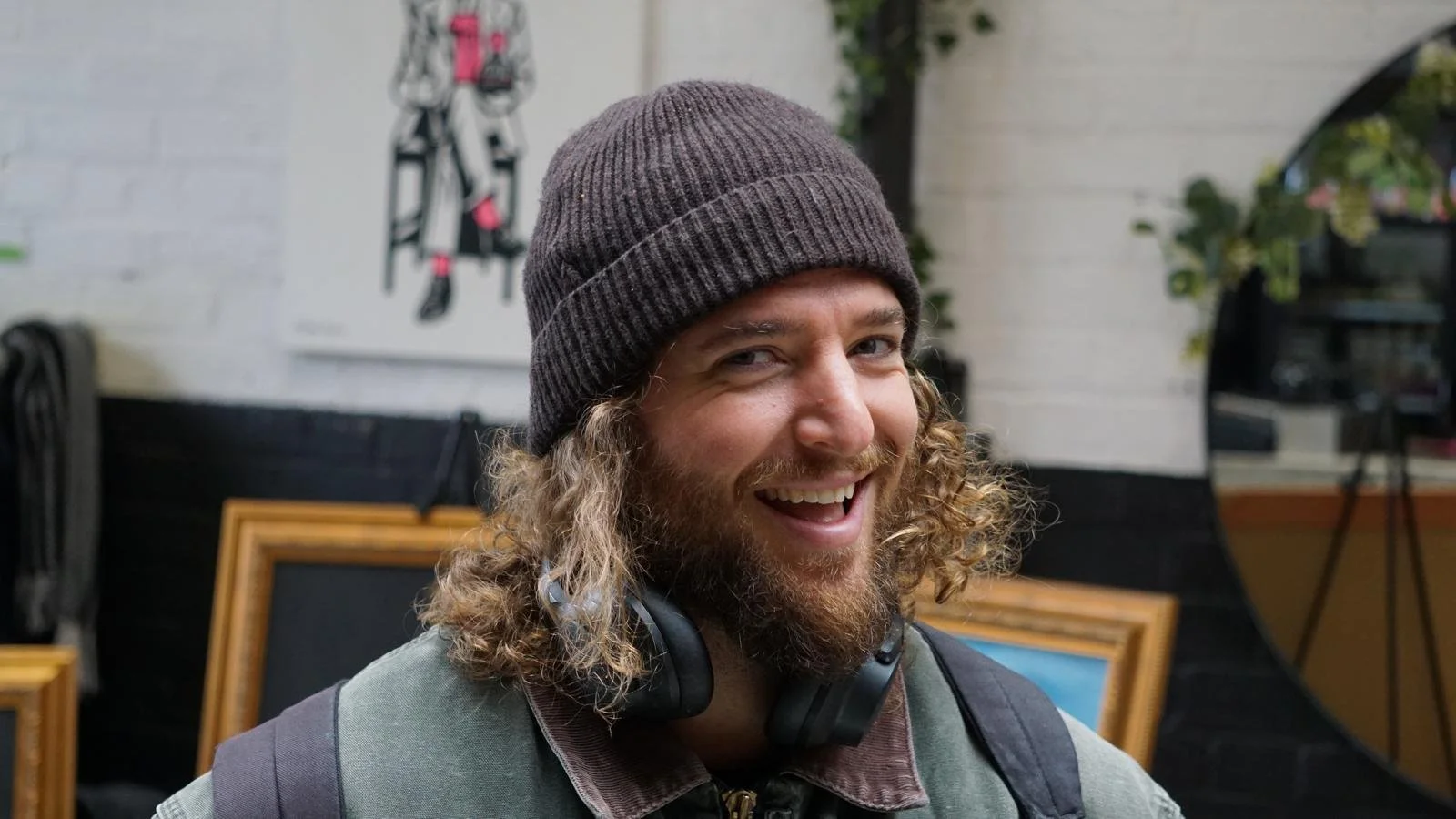 A man with curly hair, a beard, and a gray beanie, smiling with headphones around his neck, in an indoor setting with artwork and plants in the background.
