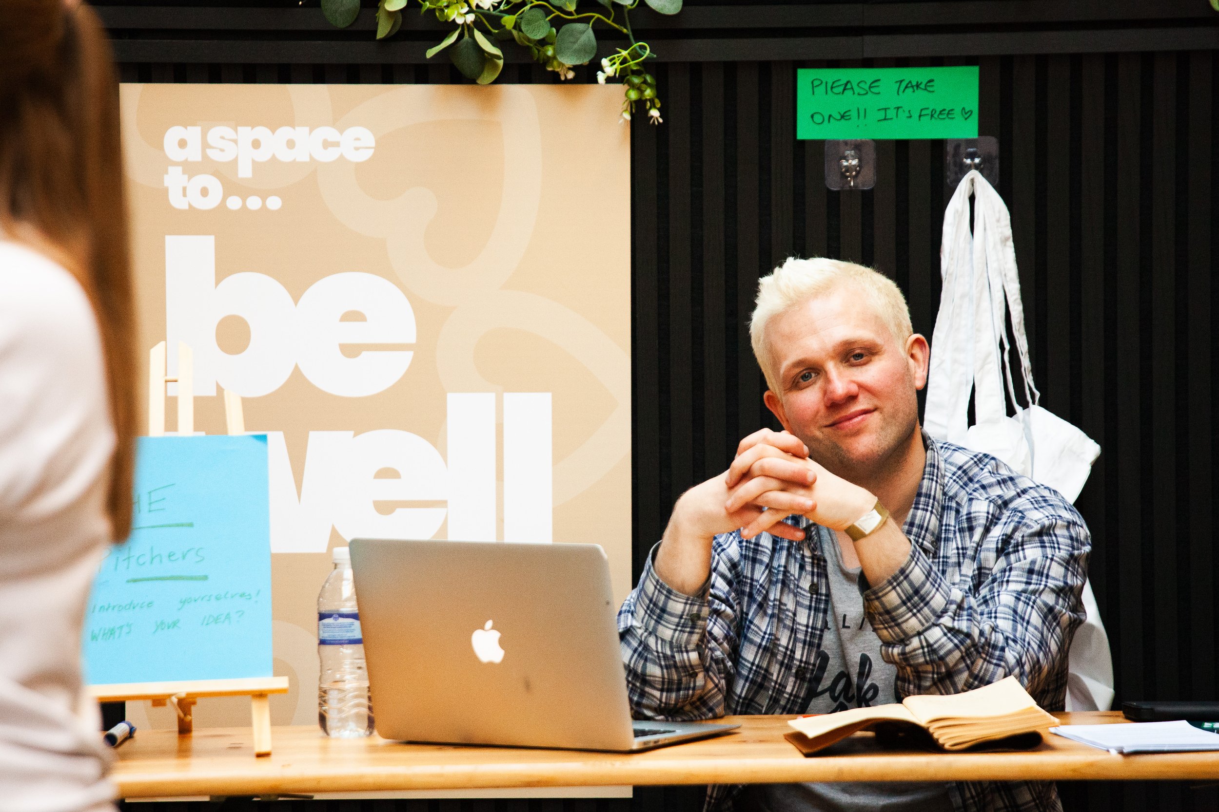 A man with blond hair sitting at a table, looking at a person outside the frame. On the table are a laptop, a water bottle, and an open notebook. Behind him, on a black wall, there are colorful signs, a greenery decoration, and some hooks with paper bags. One sign says 'a space to... be well,' and a green note says 'Please take one!! It's free.'