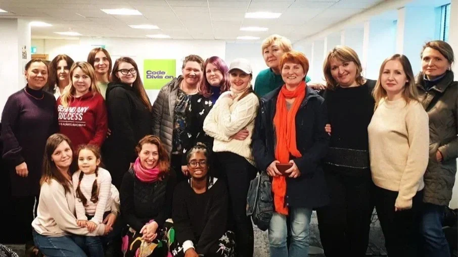 Group of women and children posing together indoors, some smiling and others with neutral expressions. Code division NomadAlba