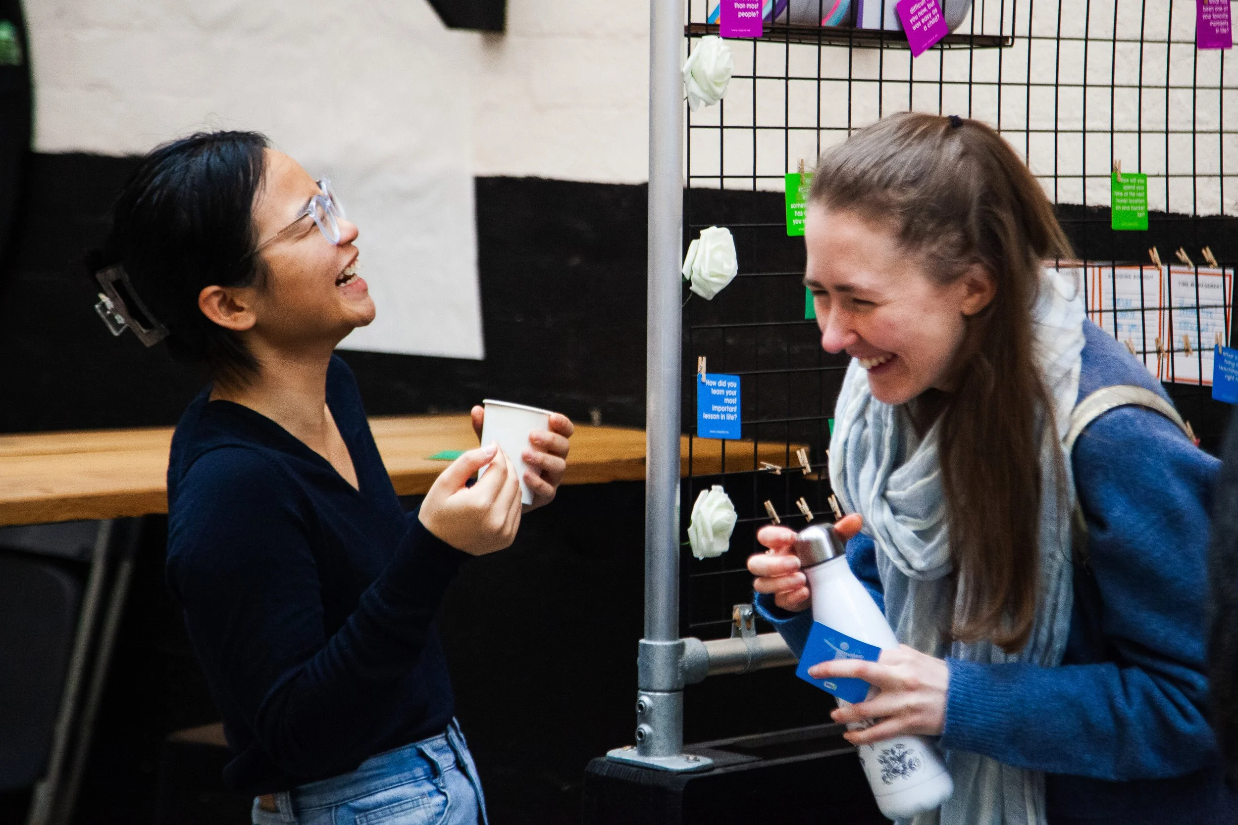 Two women are laughing and talking to each other at a social event, holding cups and a water bottle, with colorful notes and decorations in the background.