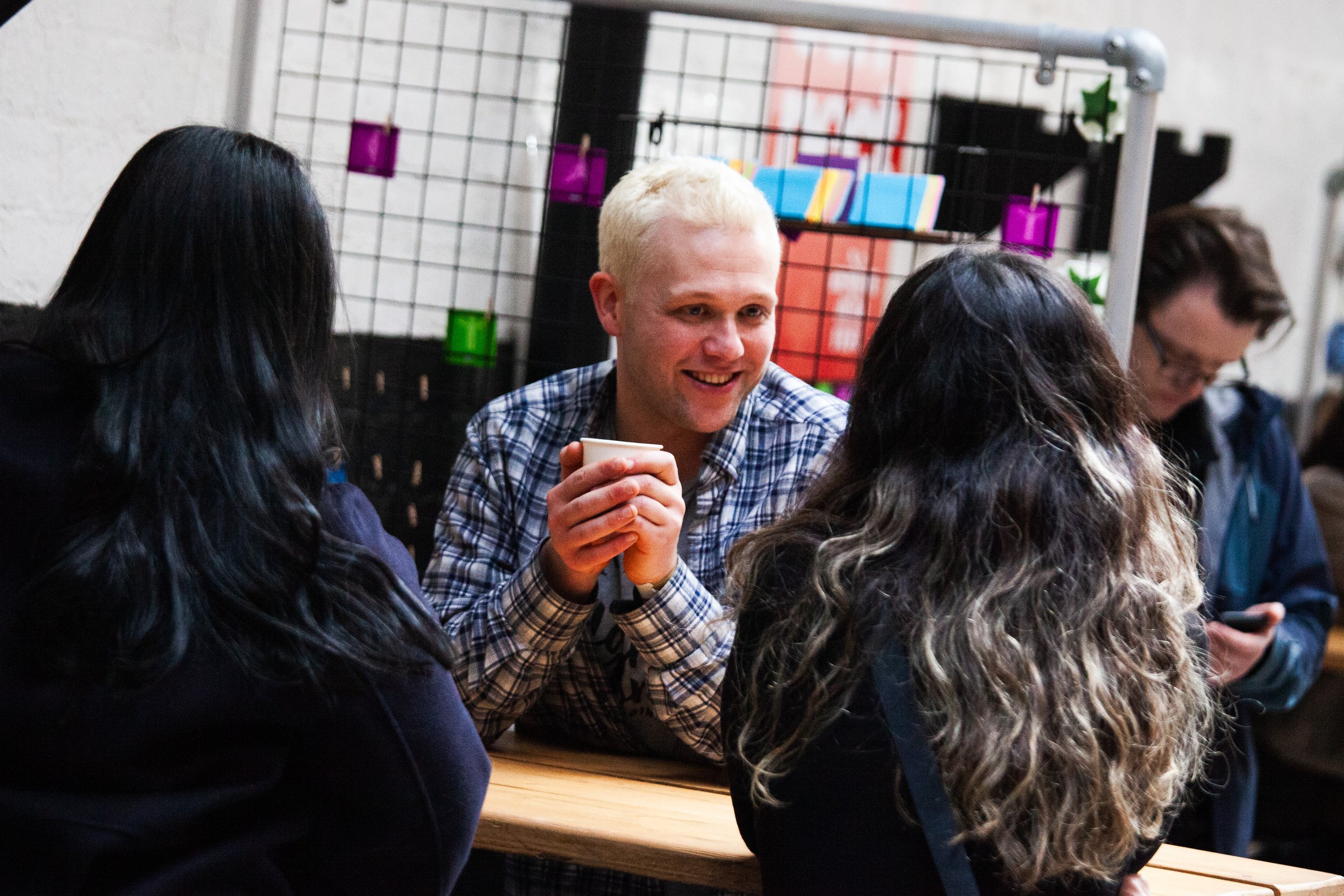 Four people sitting at a table, talking and smiling, with one holding a cup, in an indoor setting with colorful decor in the background.
