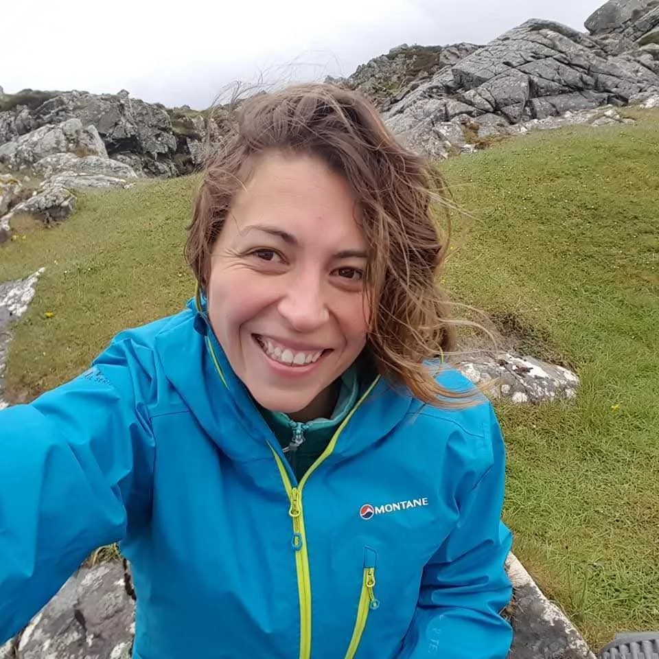 A woman takes a selfie outdoors, wearing a blue Montane jacket, with a background of rocks and grassy terrain.