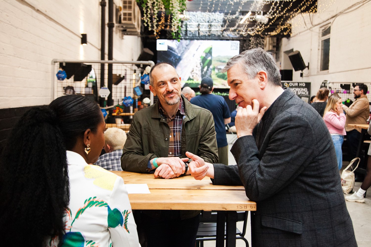 Three people having a conversation at a social gathering or event, with a woman and two men, one with gray hair and the other with a shaved head. The setting is indoors with decorative string lights and a large screen in the background.