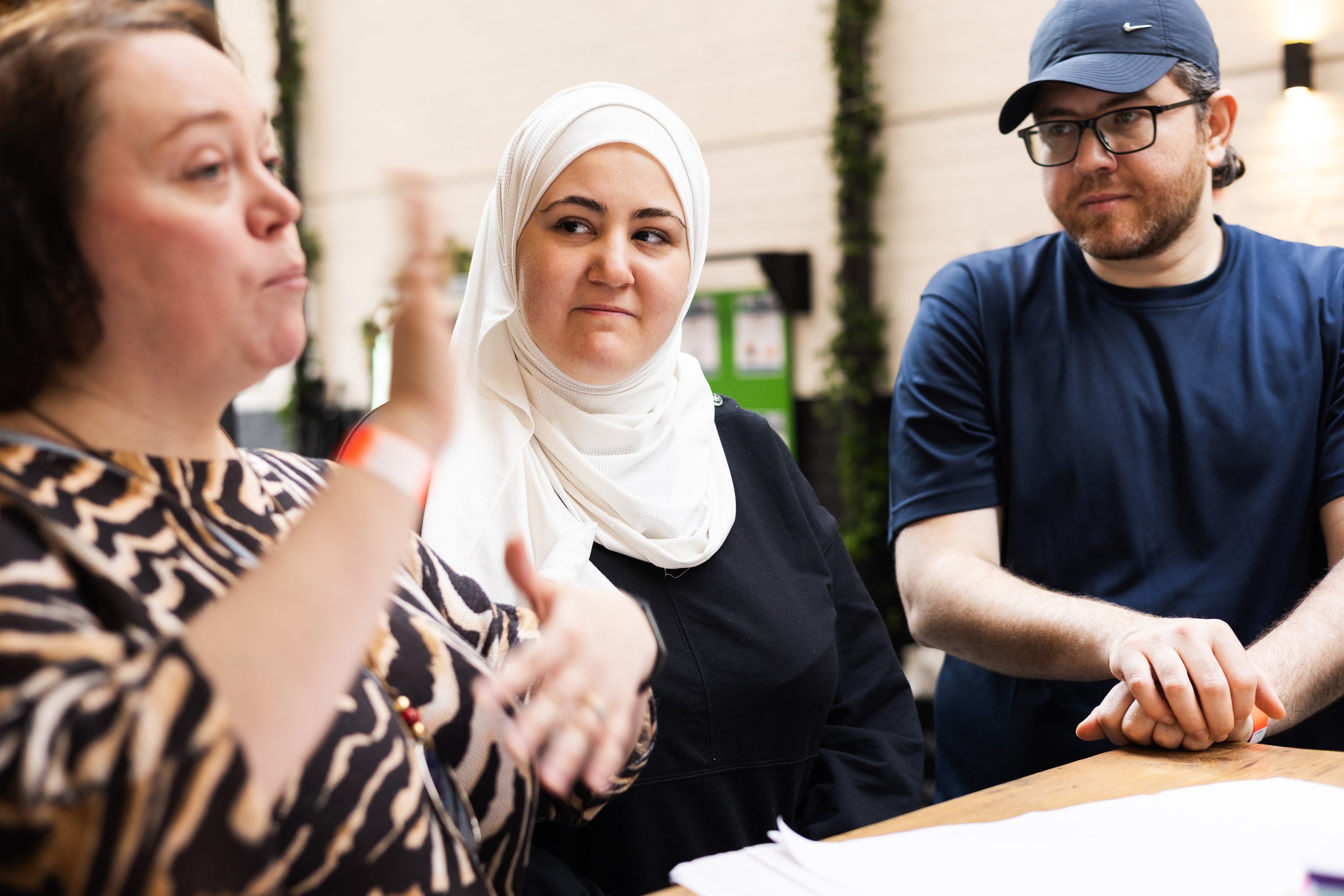 Three people engaged in conversation indoors, with one woman speaking, a woman wearing a white hijab listening, and a man in a blue cap and glasses looking on.