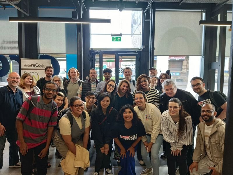 Group photo of diverse people inside a modern building with large windows and sunlight coming in.