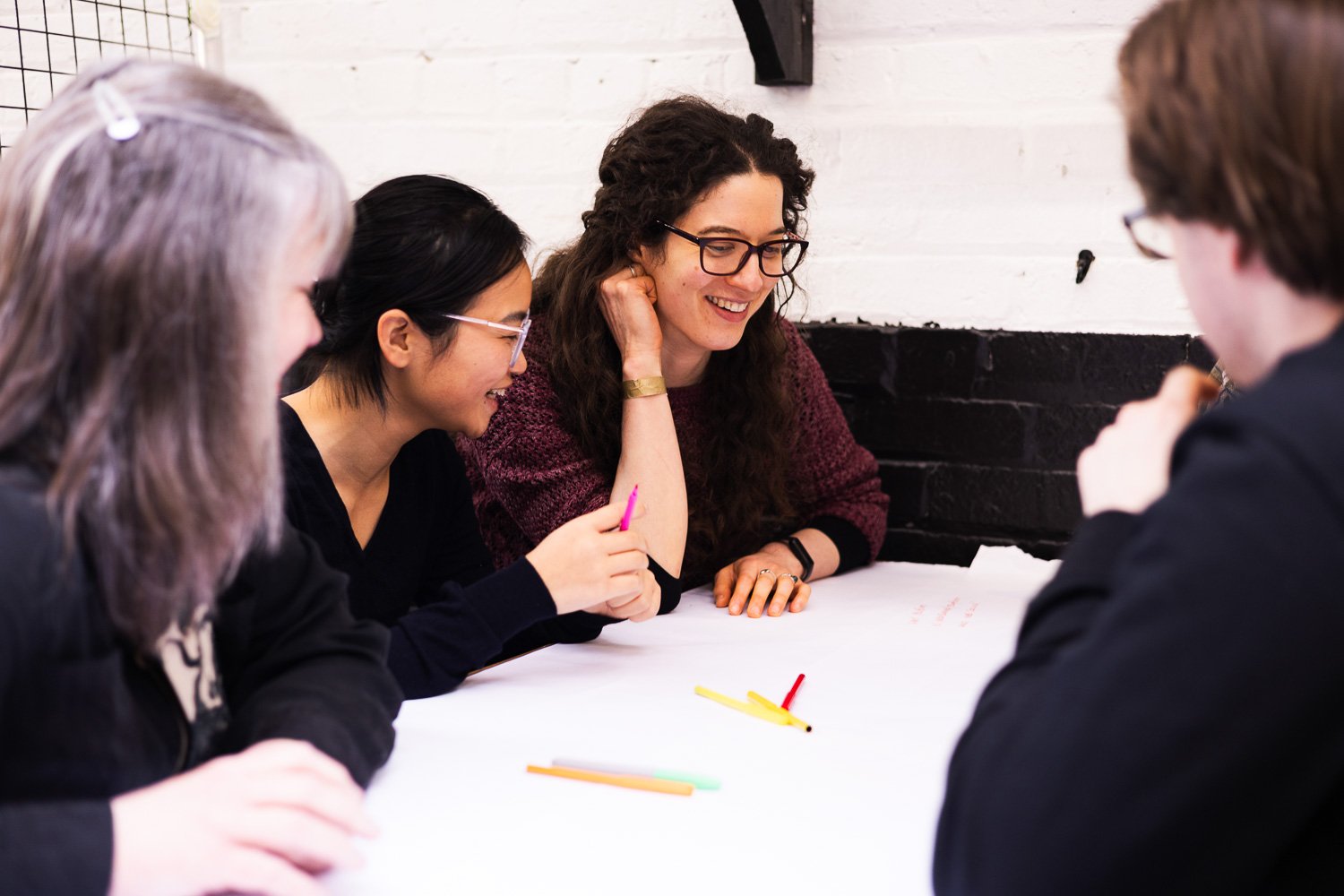 Four people sitting around a table, smiling and talking, with colorful pens and paper in front of them, in a room with white brick walls.