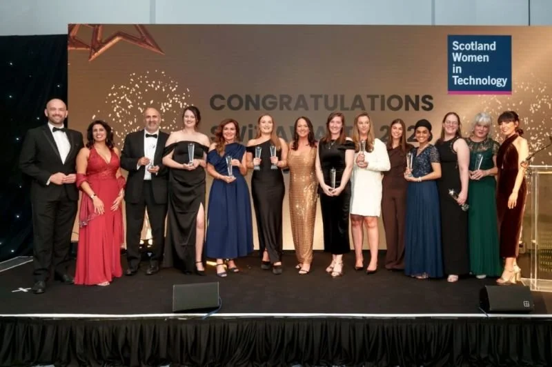 Group of women and men in formal attire celebrating at an event for Scotland Women in Technology, holding awards, on a stage with a large congratulatory sign and decorations.