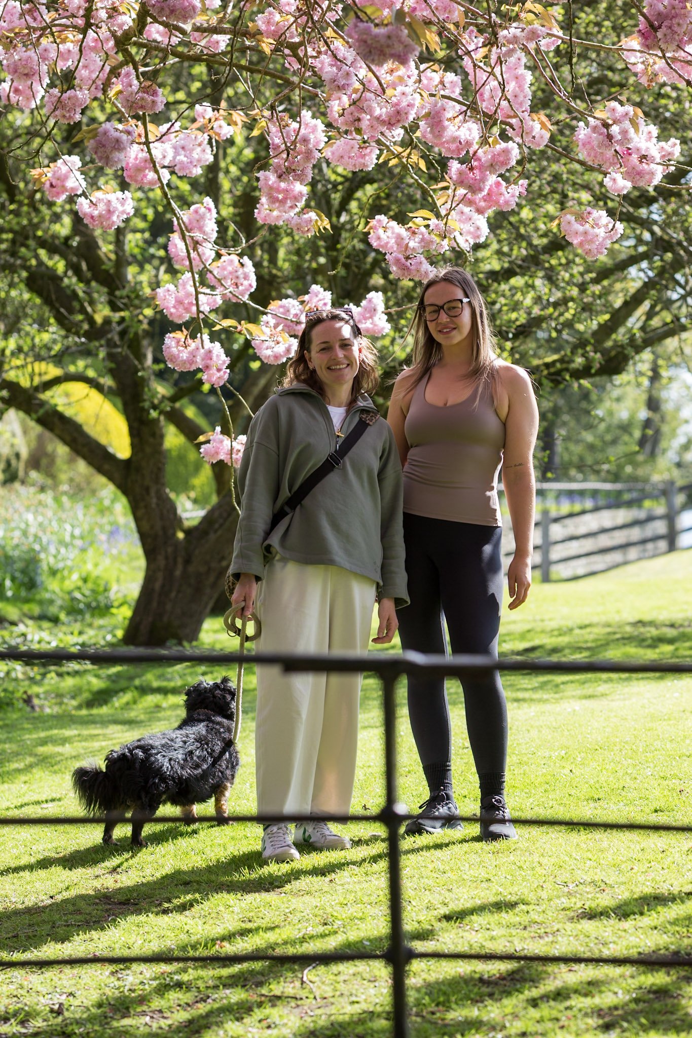 Two women standing outdoors in front of a blooming pink cherry blossom tree. One woman has long hair, glasses, and is wearing a tank top and leggings. The other woman has shoulder-length hair, is smiling, wearing a jacket, and holding a leash with a dog. The dog is black and gray, sitting on the grass facing away from the camera.