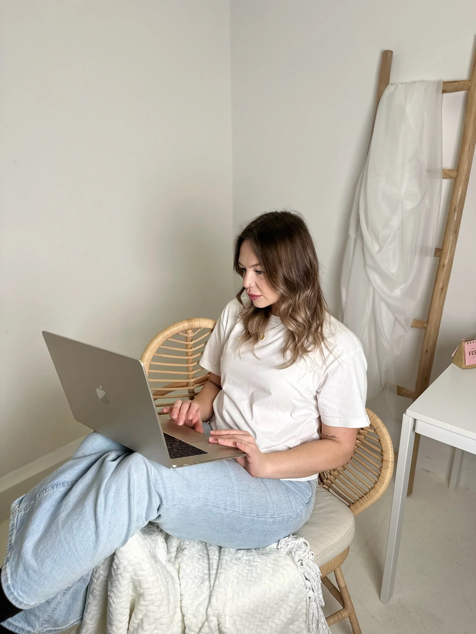 A woman with brown hair sits on a beige cushioned chair, using a silver laptop on her lap. Another person, wearing light blue jeans and a light-colored blanket, is sitting with their legs up on the chair, partially visible in the foreground. The background shows a plain white wall, a wooden ladder leaning against it with a white cloth draped over one of the rungs, and a white side table with a pink box labeled 'FEED'.