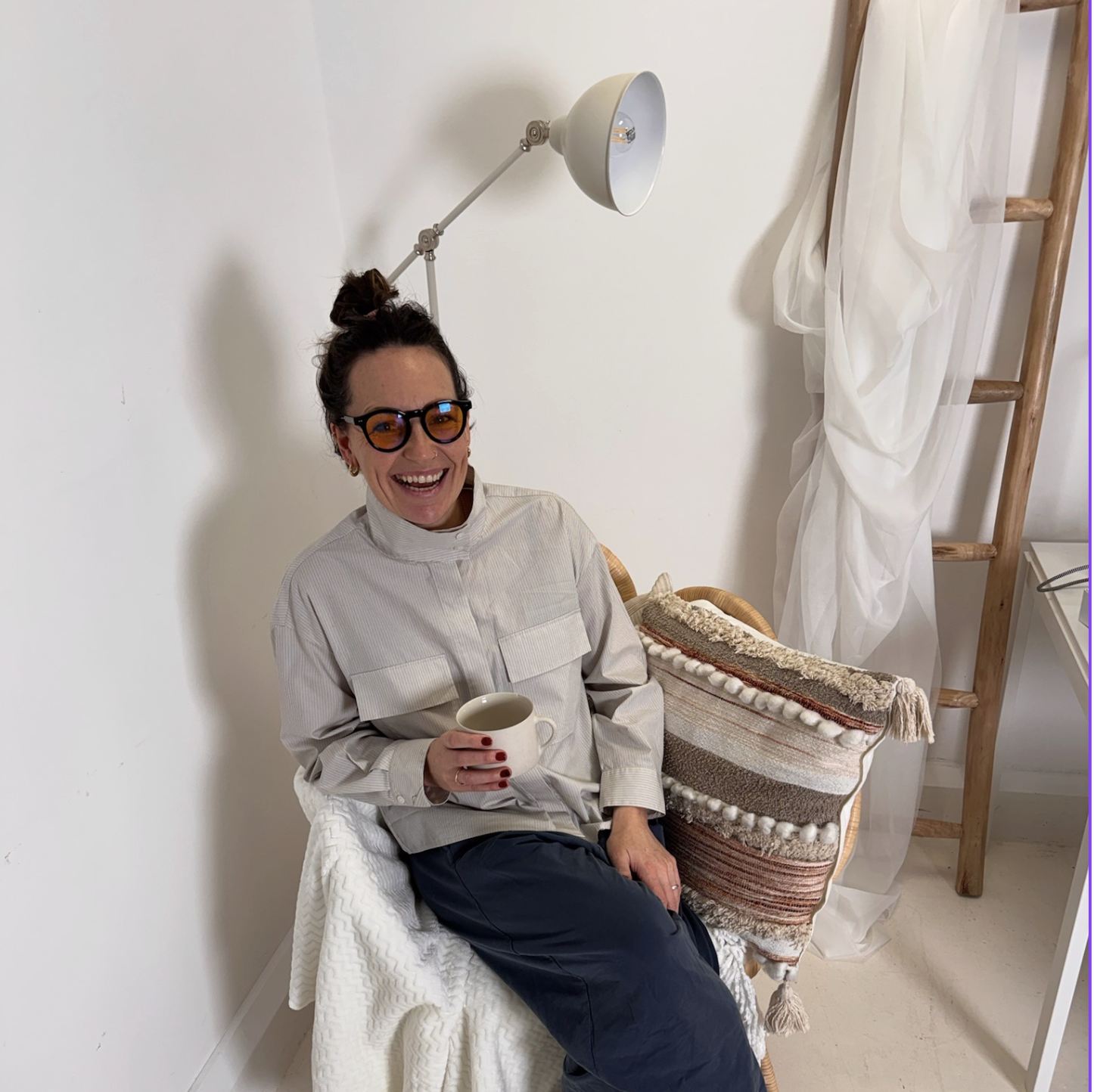 A woman with dark hair in a bun, wearing glasses and a beige button-up shirt, smiling, sitting on a chair and holding a white mug. Behind her, a white wall, a wooden ladder with draped white fabric, and a woven basket with various patterns and textures are visible.