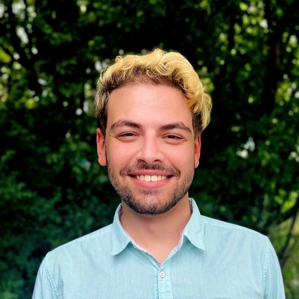 A young man with blond and brown hair smiling outdoors with green trees in the background.