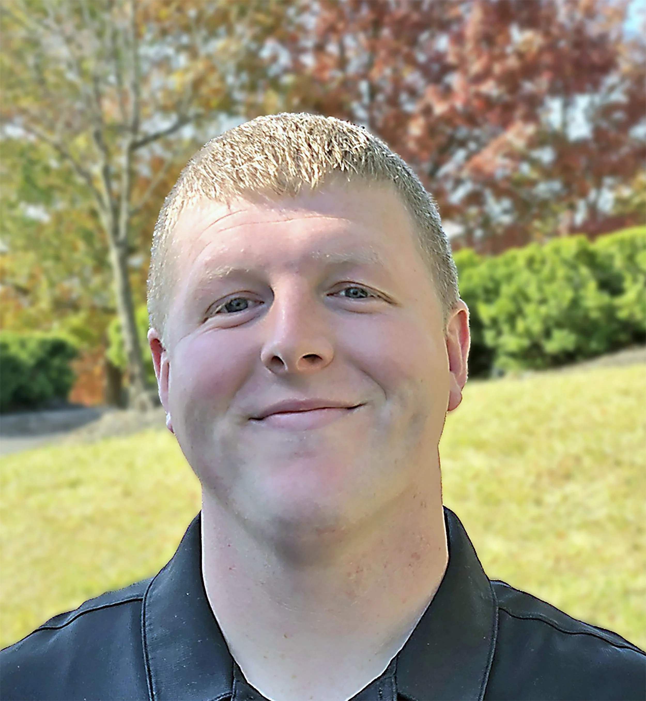 A young man with short blonde hair and a slight smile, wearing a dark leather jacket, standing outdoors in a park with colorful autumn trees and green bushes in the background.
