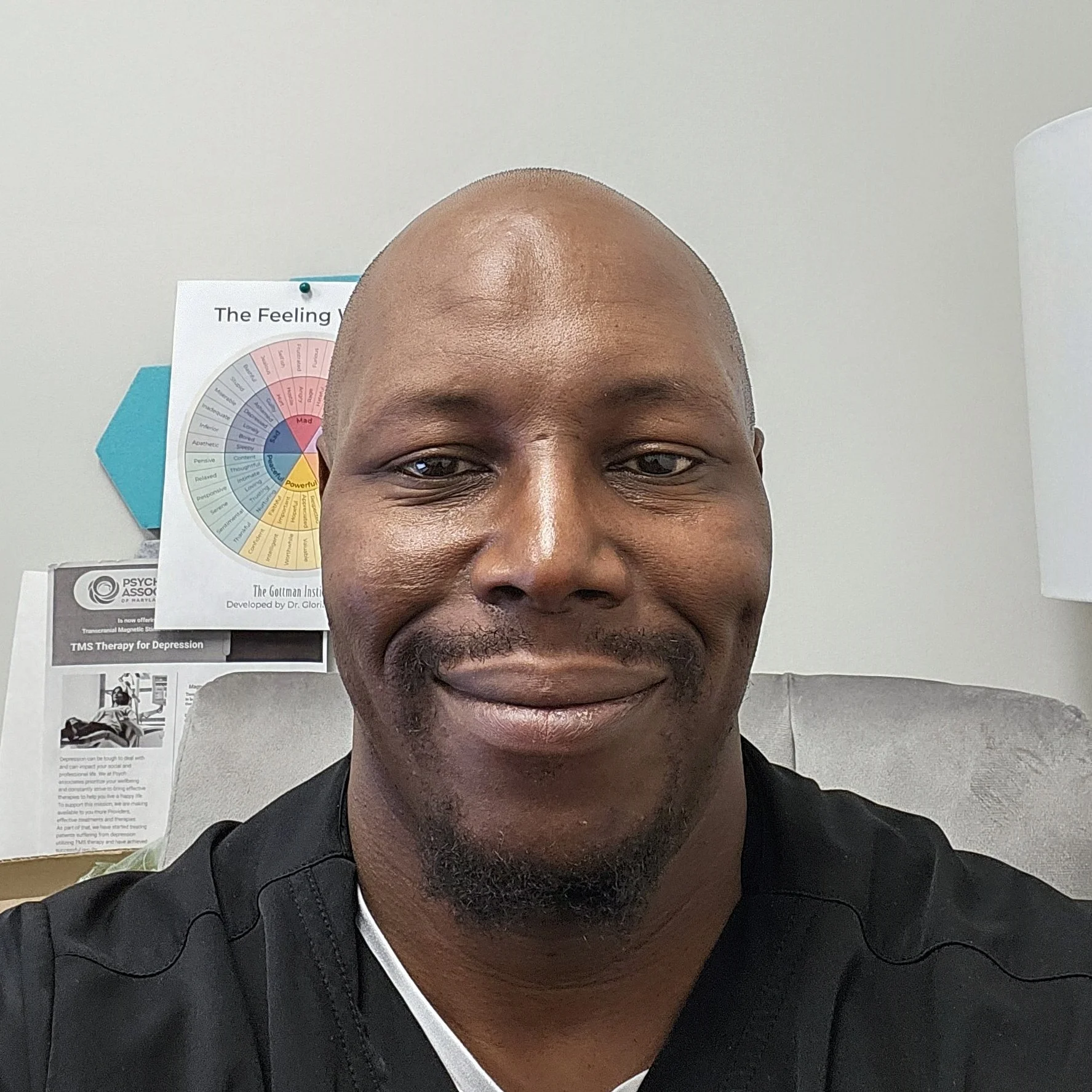 Close-up of a smiling African American man with a bald head, seated in an office with psychological posters on the wall behind him.