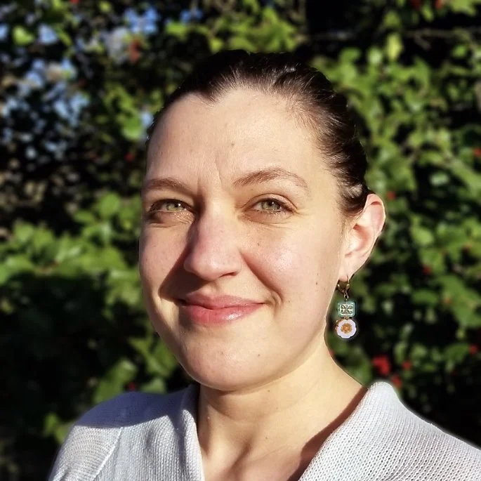 A woman with fair skin, brown hair pulled back, and earrings, smiling outdoors with green foliage in the background.