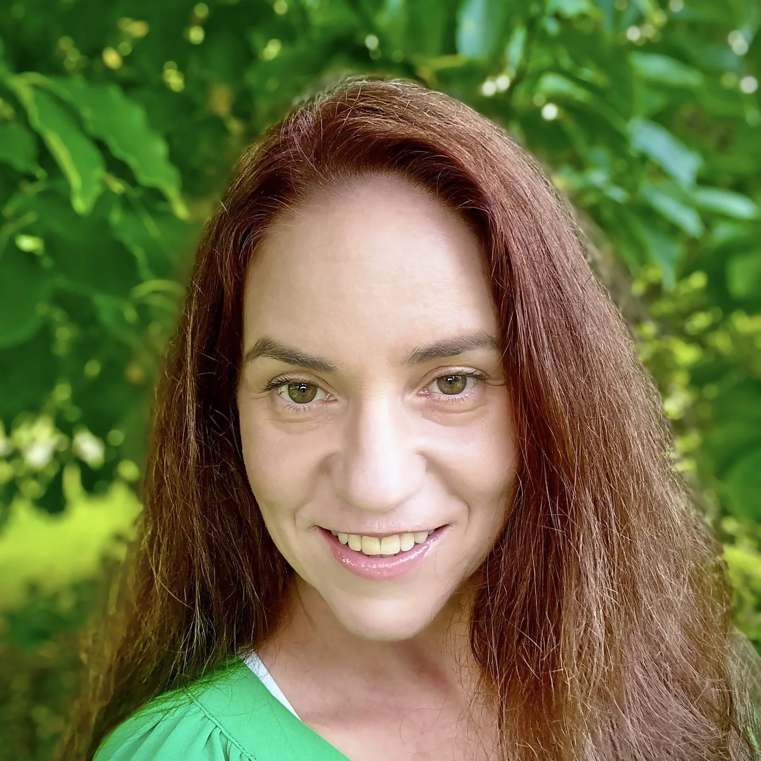 Close-up of a smiling woman with long reddish-brown hair and green eyes, wearing a green top outdoors with green foliage in the background.
