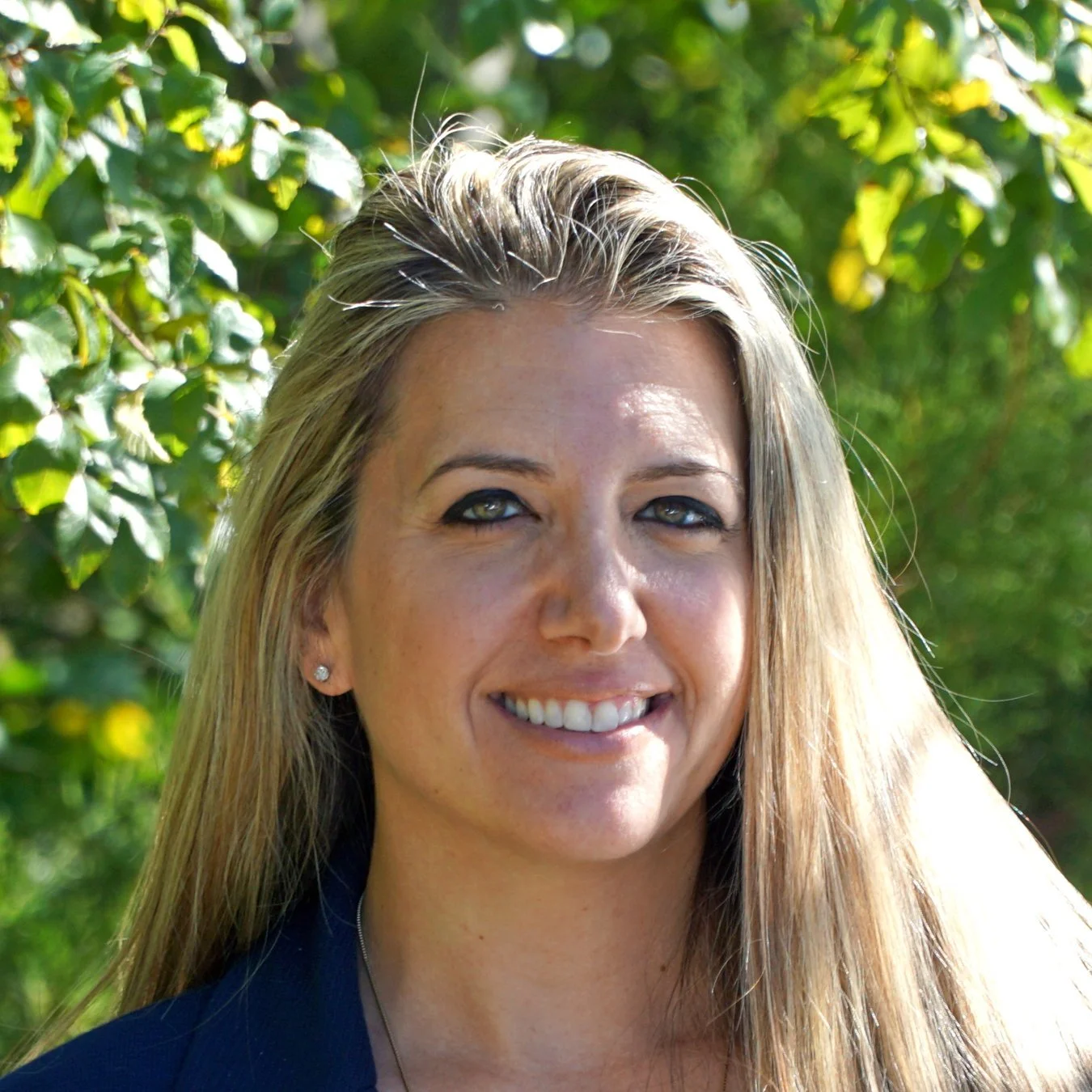 Close-up of a smiling woman with long blonde hair and blue eyes, outdoors with green foliage in the background.