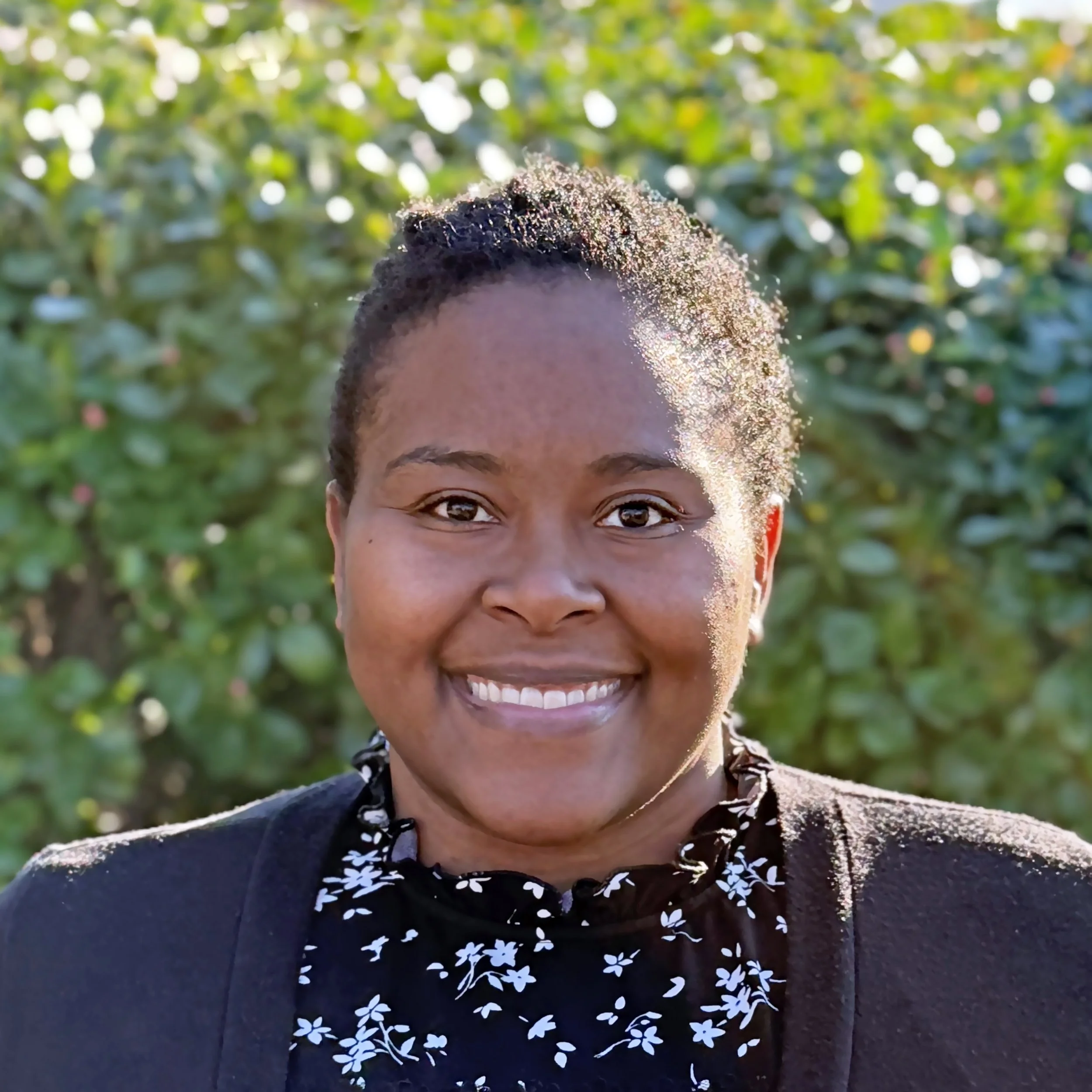 Close-up of a smiling woman with short, curly hair outdoors, wearing a black cardigan over a floral blouse, with green foliage in the background.