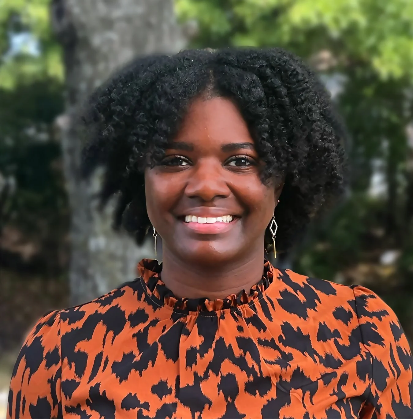 A smiling Black woman with short curly hair, wearing a leopard print blouse and drop earrings, standing outdoors with a tree and greenery in the background.