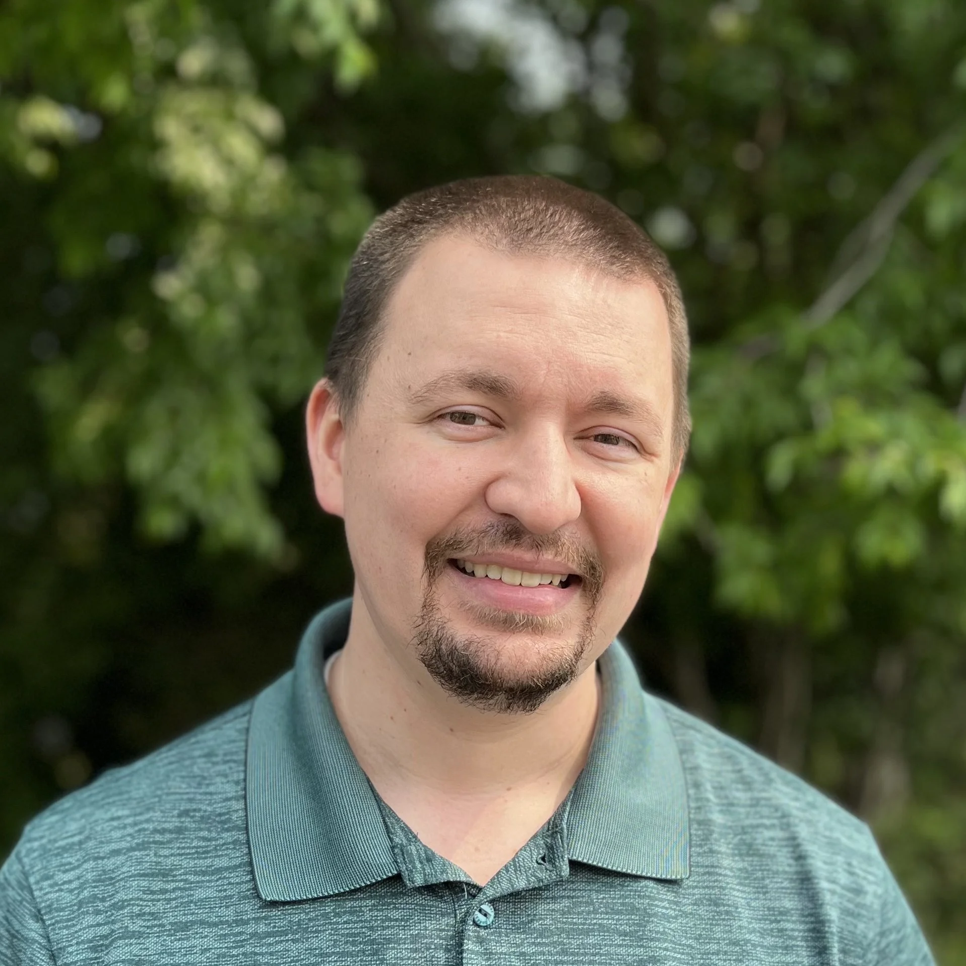 A smiling man with short brown hair, a mustache and goatee, wearing a teal polo shirt, standing outdoors with green foliage in the background.