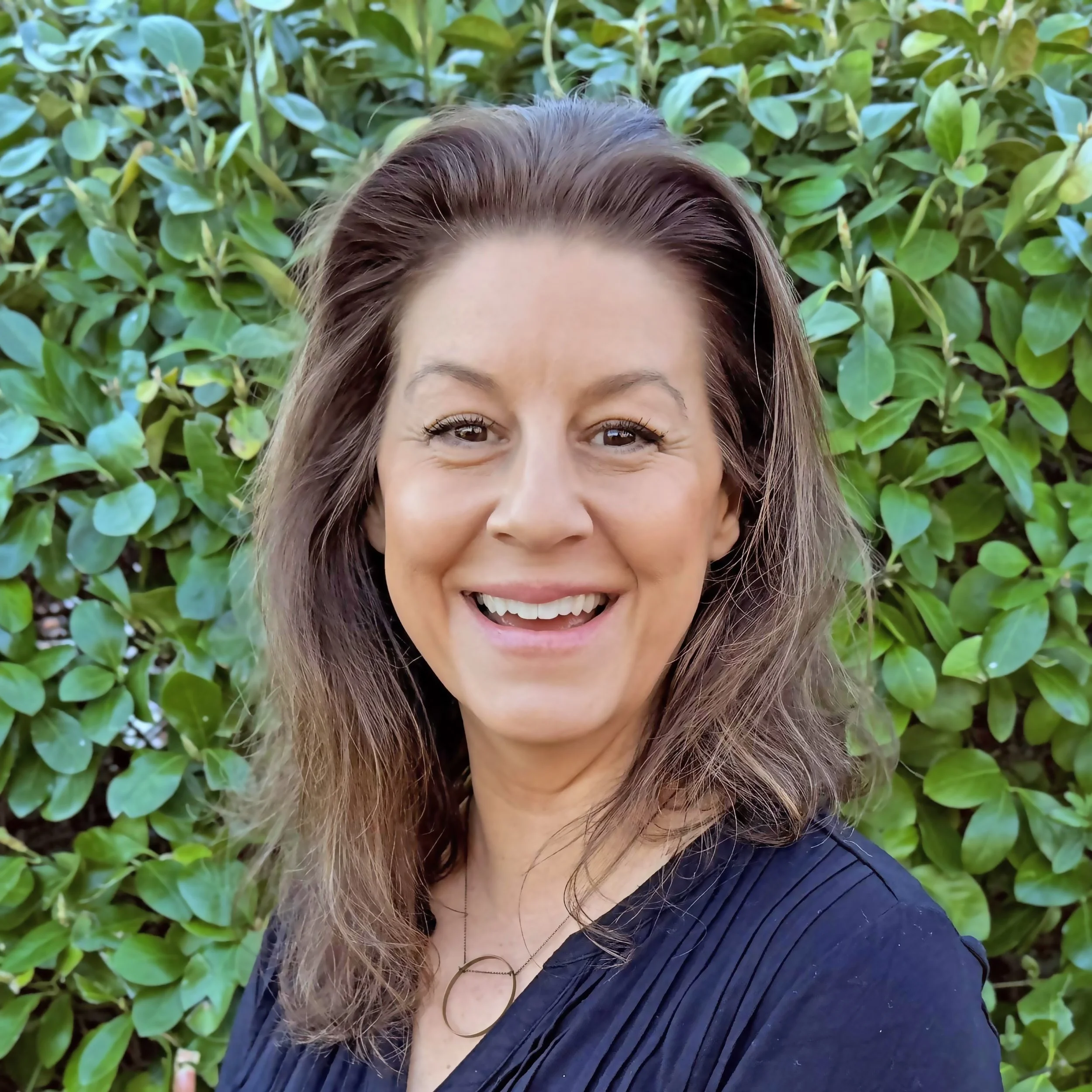 A woman smiling outdoors with green leafy background.