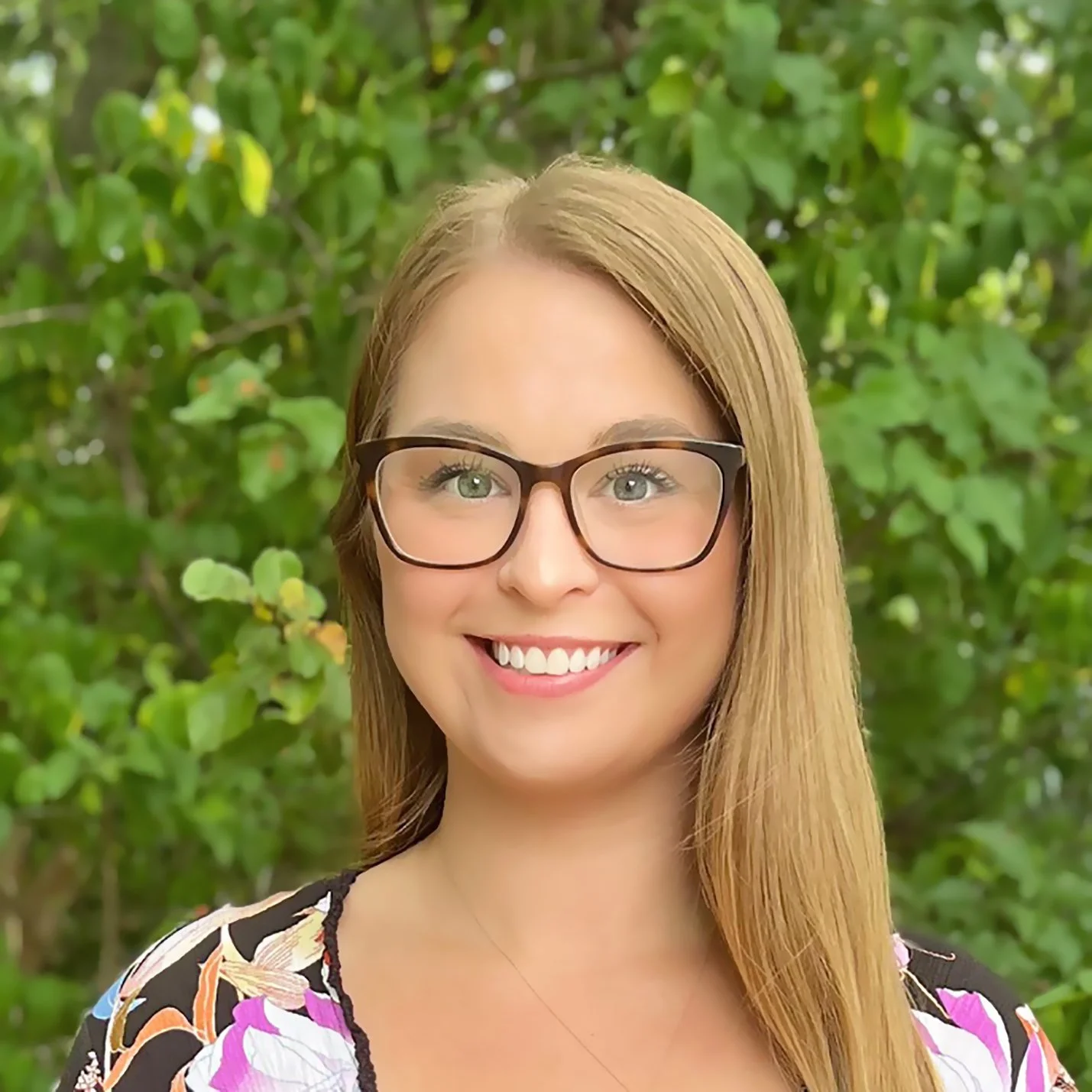 A woman with long, straight blonde hair and glasses smiling outdoors in front of green leafy foliage.