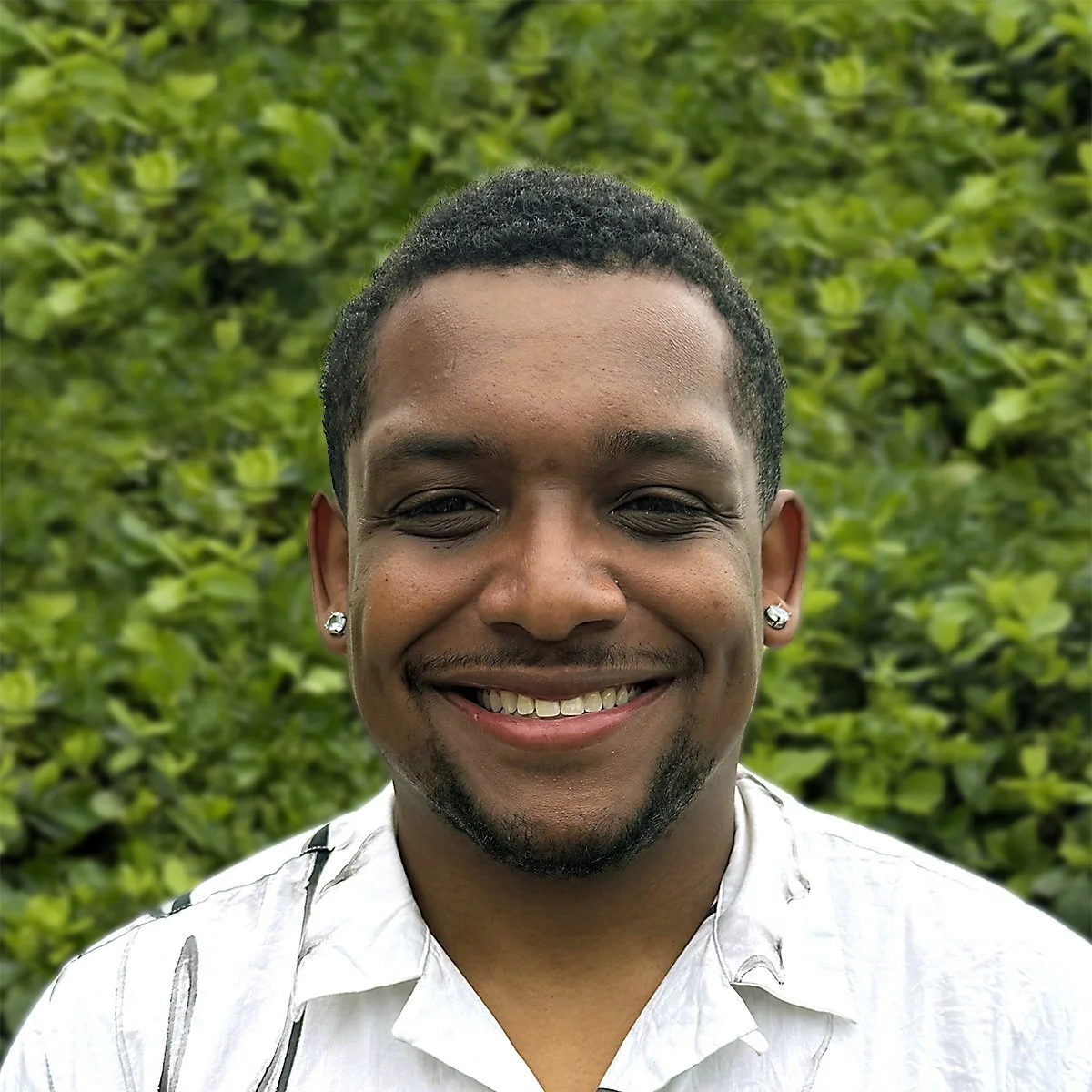 Portrait of a smiling young man with short hair, earrings, and a white collared shirt, standing outdoors in front of green foliage.