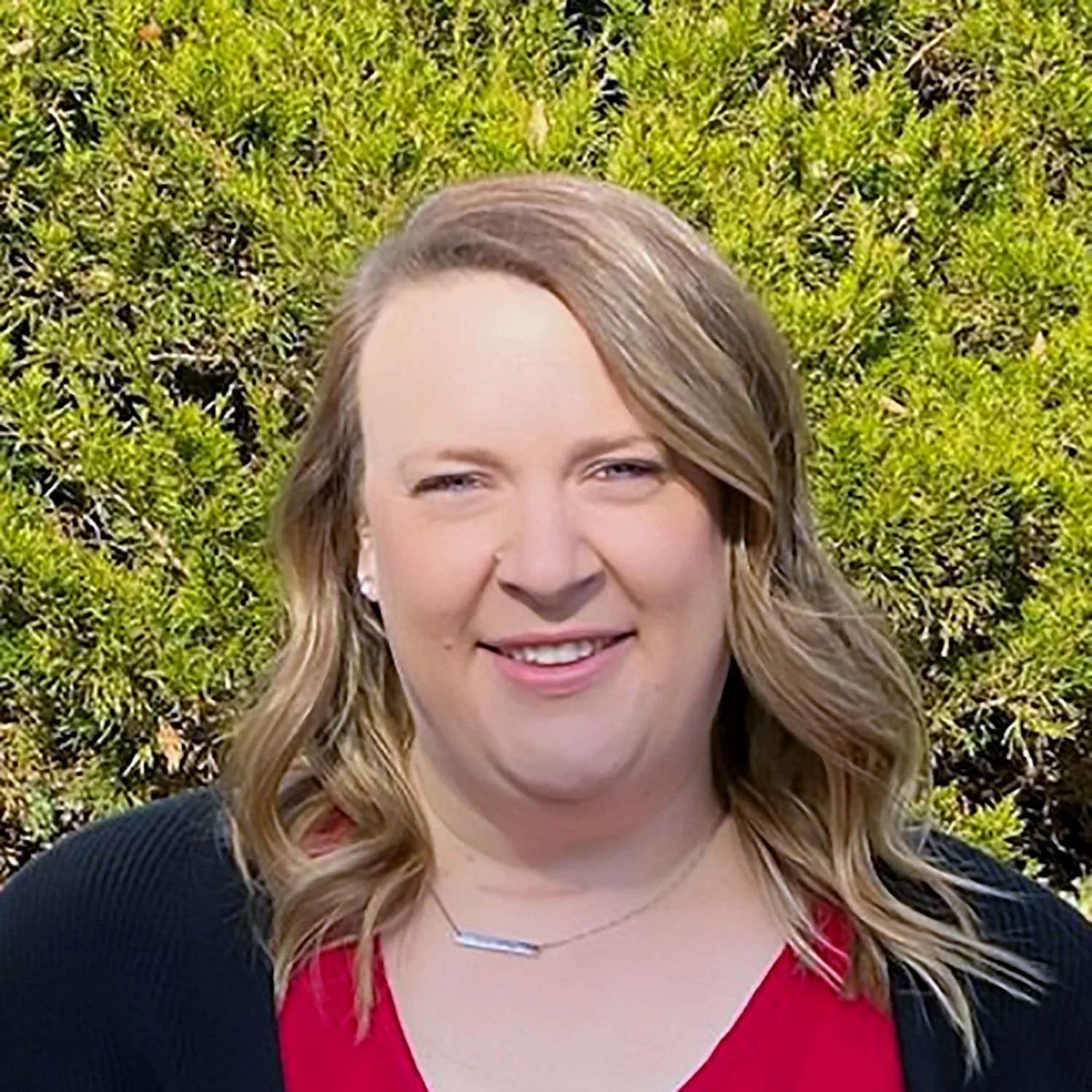 A woman with wavy blonde hair smiling outdoors with a leafy green hedge in the background.