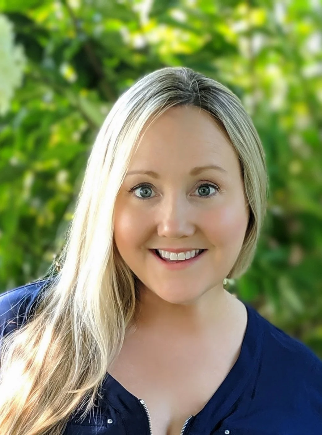 A smiling woman with long blonde hair and blue eyes, wearing a navy blue shirt, outdoors with green foliage in the background.