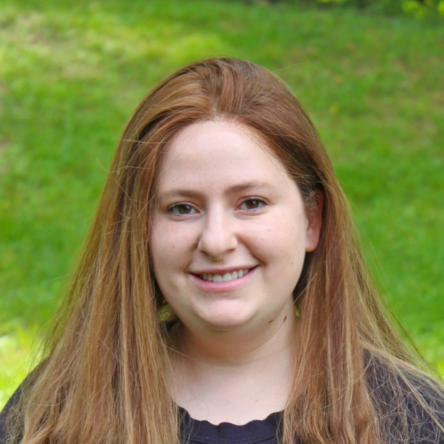 A young woman with long red hair smiling outdoors in front of a green grassy background.