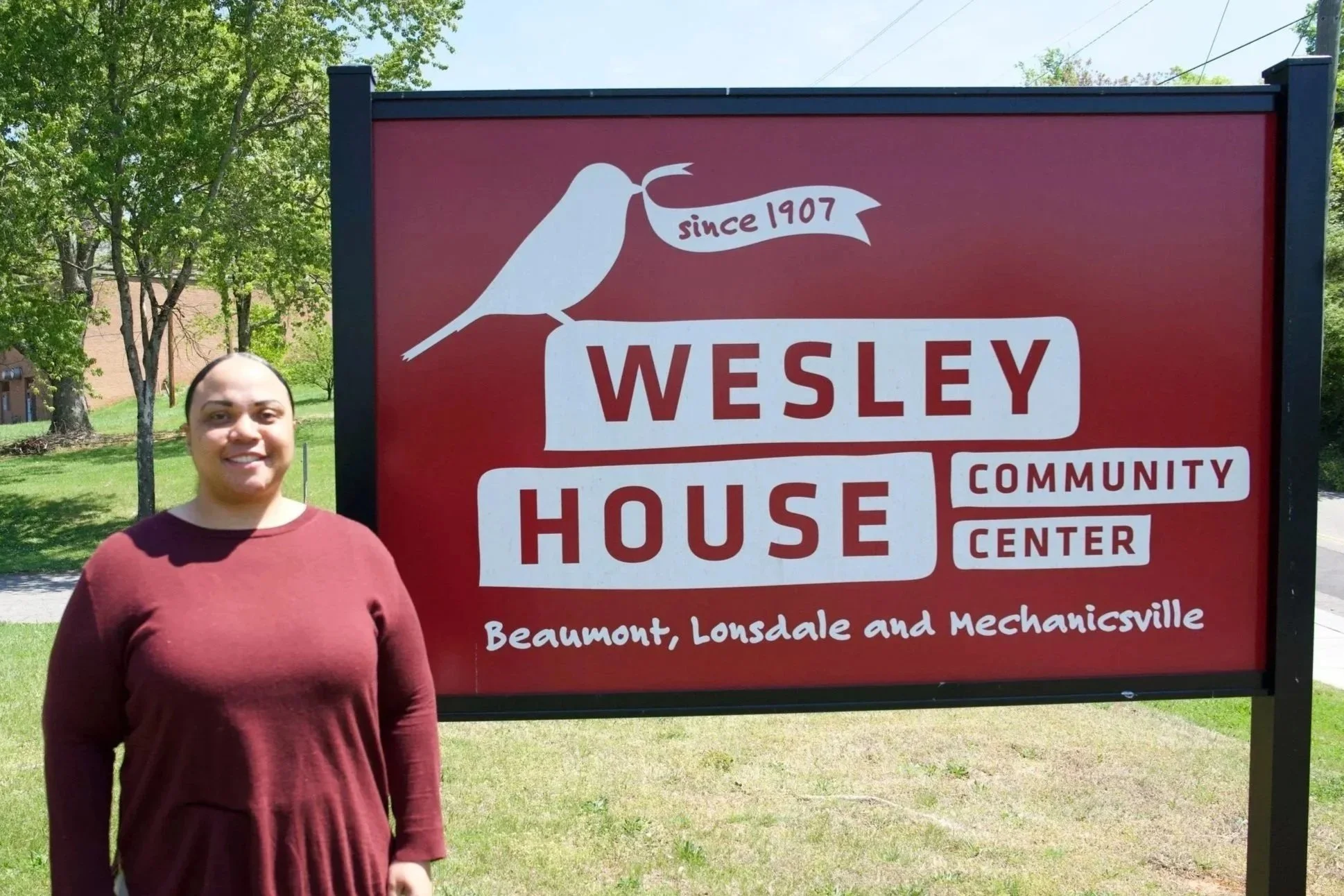A picture of a woman in front of the Wesley House sign. Her shirt is red and her pants are brown.