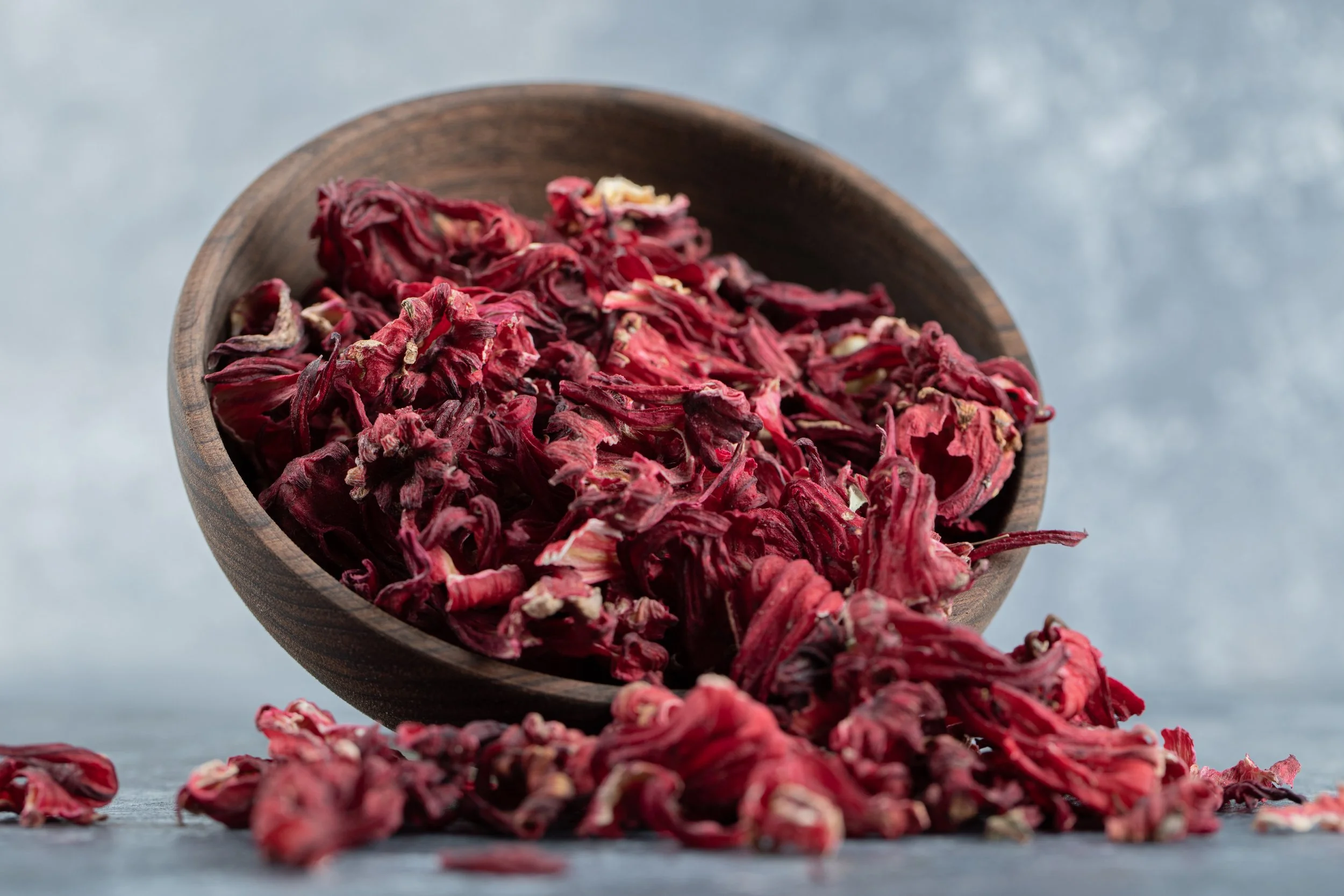 Dried hibiscus petals in a wooden bowl with some spilled on a gray surface.