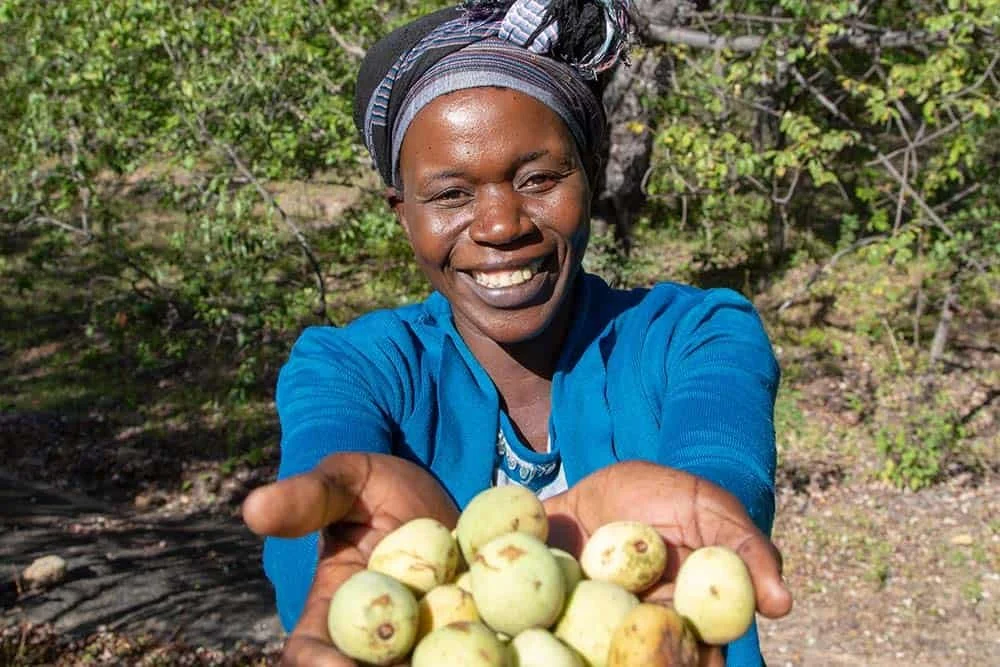 A woman smiling and holding out a handful of small, round, light green fruits outdoors in a natural setting with trees and bushes.