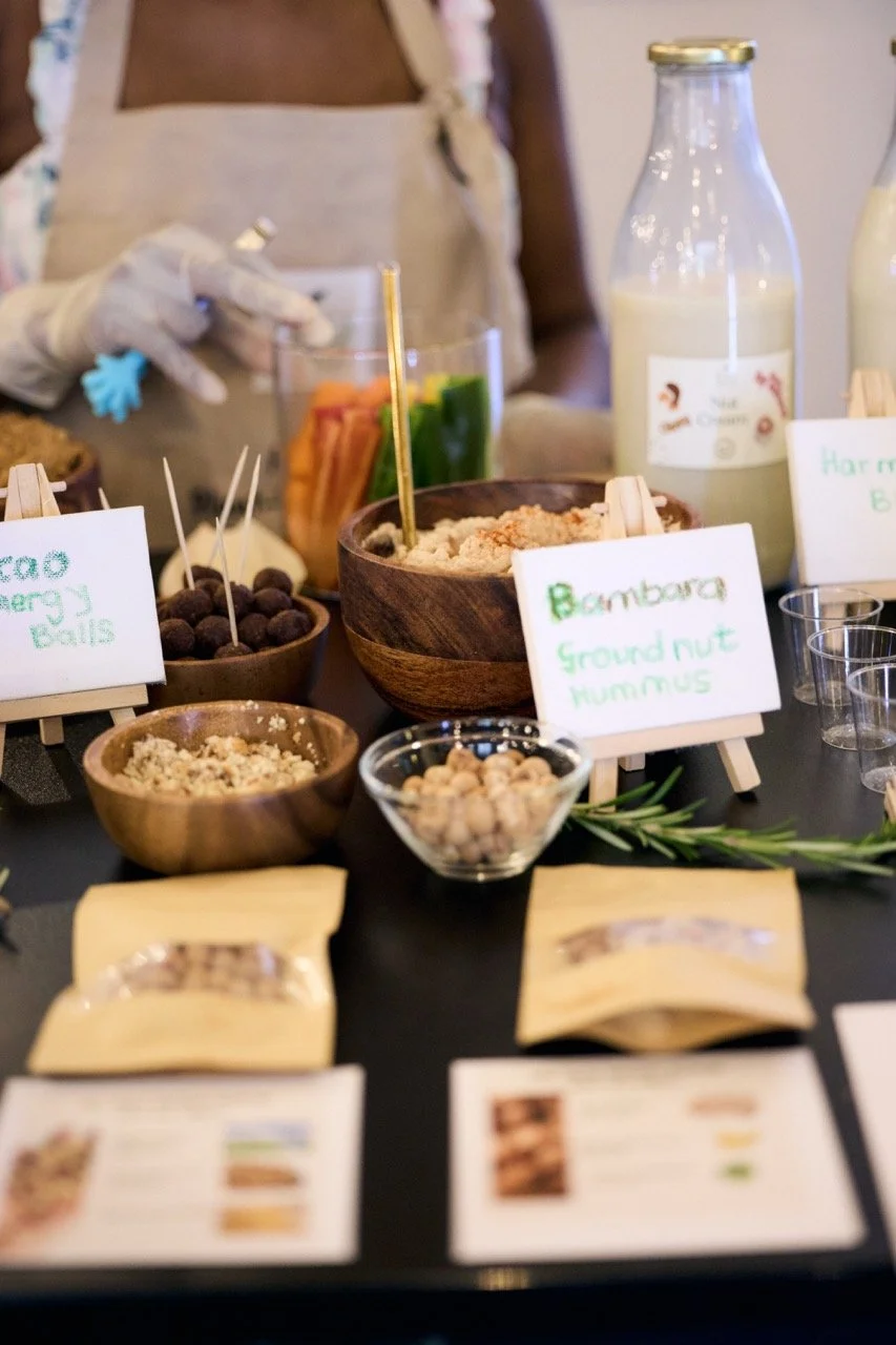 Assorted natural food products on a table, including bowls of ground nuts, chickpeas, and hummus, with a person in the background wearing gloves and an apron, preparing food.