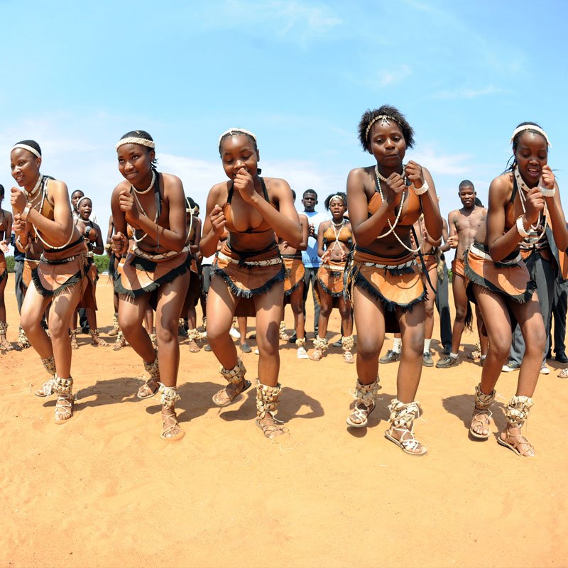 Group of young women in traditional dance outfits performing a dance on the dirt ground outdoors under a clear blue sky.
