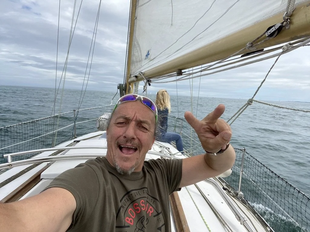 Un homme souriant prend un selfie sur un bateau à voile en mer, avec une femme assise en arrière, regardant au loin, par un temps nuageux.