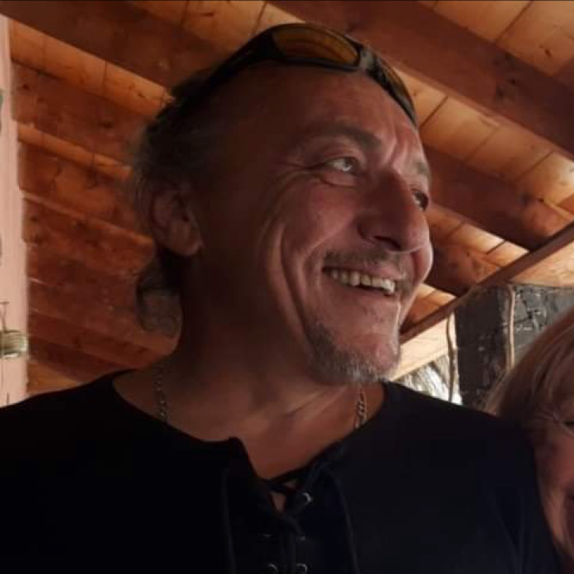 Un homme souriant portant des lunettes de soleil sur la tête, avec une barbe grise, dans un intérieur en bois.