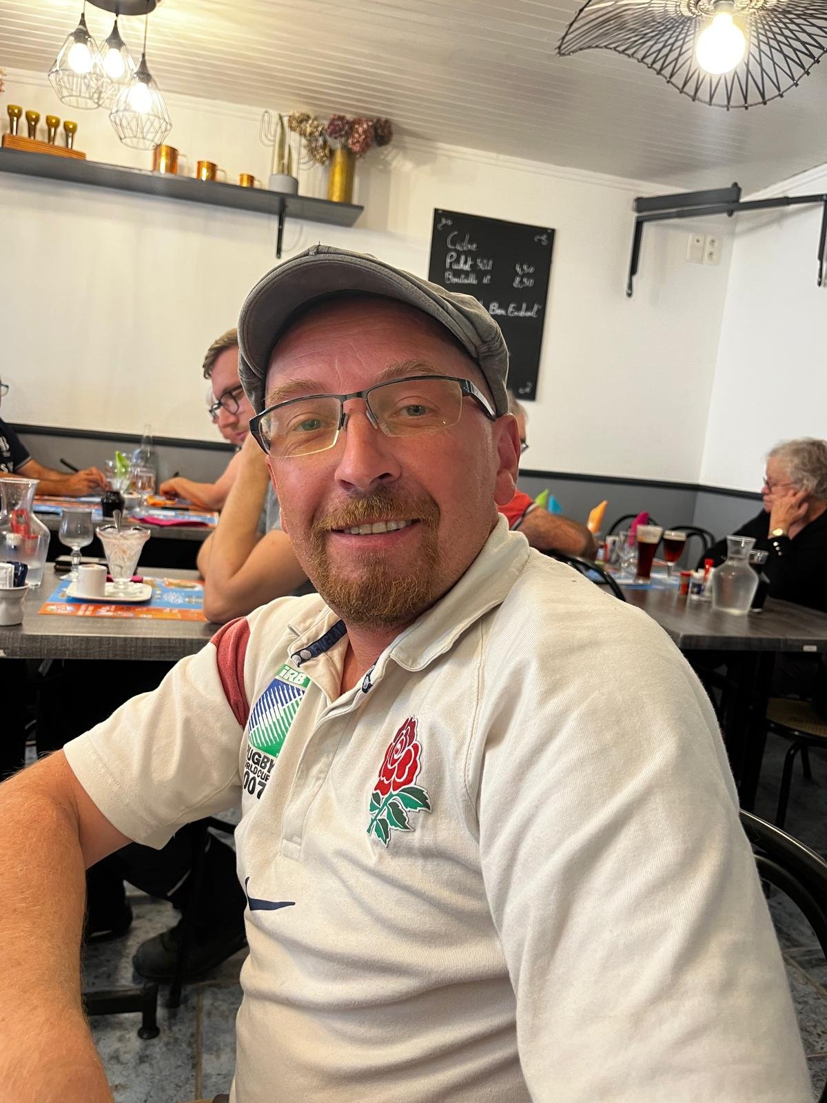Un homme souriant portant une casquette, des lunettes et un maillot blanc avec des logos, assis dans un restaurant avec d'autres personnes en arrière-plan.