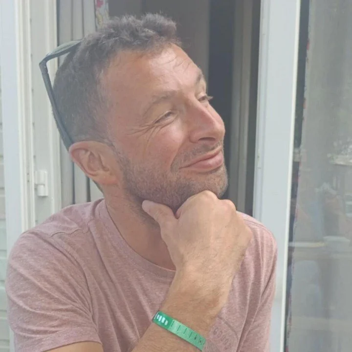 Un homme souriant, avec des lunettes sur la tête, portant un t-shirt rose, assis près d'une fenêtre, se reposant sur sa main.