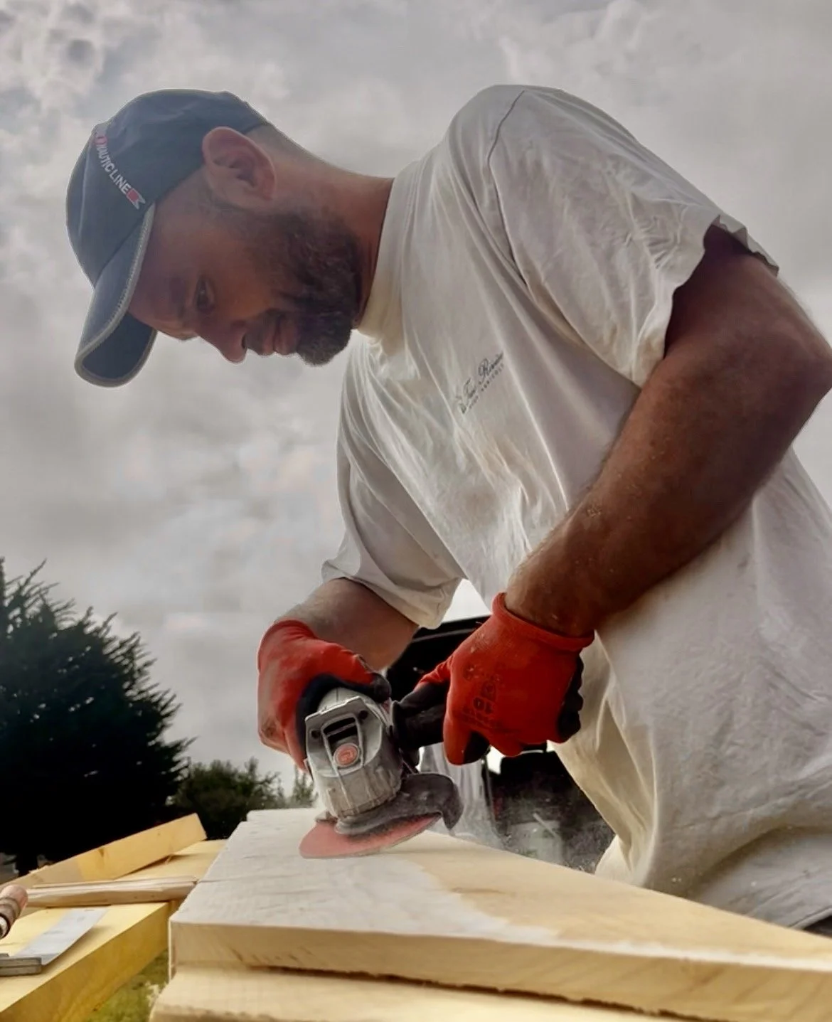 Un homme coupe du bois à l'extérieur avec une scie électrique, portant des gants orange et un chapeau bleu, avec un ciel nuageux en arrière-plan.
