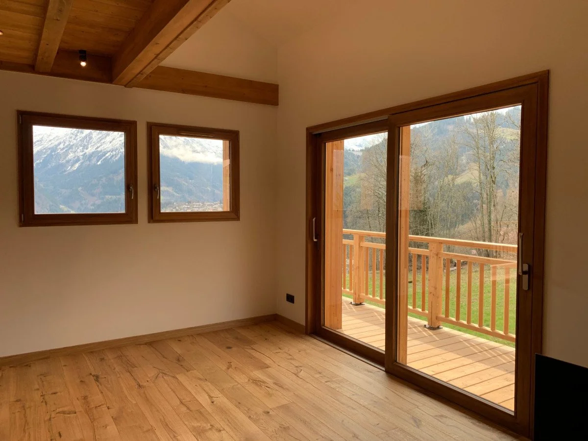 Chambre avec deux fenêtres et une porte-fenêtre donnant sur un balcon en bois, avec vue sur des montagnes et des arbres.