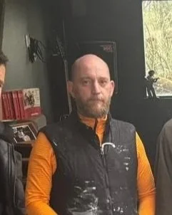 Homme chauve avec barbe portant un gilet noir et un t-shirt orange, assis à l'intérieur, avec une fenêtre laissant entrer de la lumière naturelle.