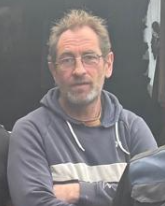 Homme portant des lunettes, cheveux grisonnants, porte un pull gris avec des rayures, croise les bras, debout devant un fond sombre.