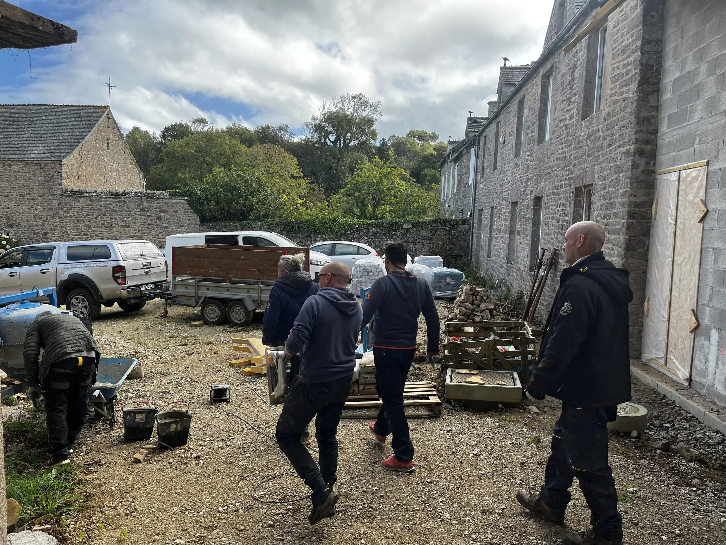 Groupe de personnes travaillant sur un chantier de construction ou de rénovation à l'extérieur d'un bâtiment en pierre, avec divers outils et matériaux autour.