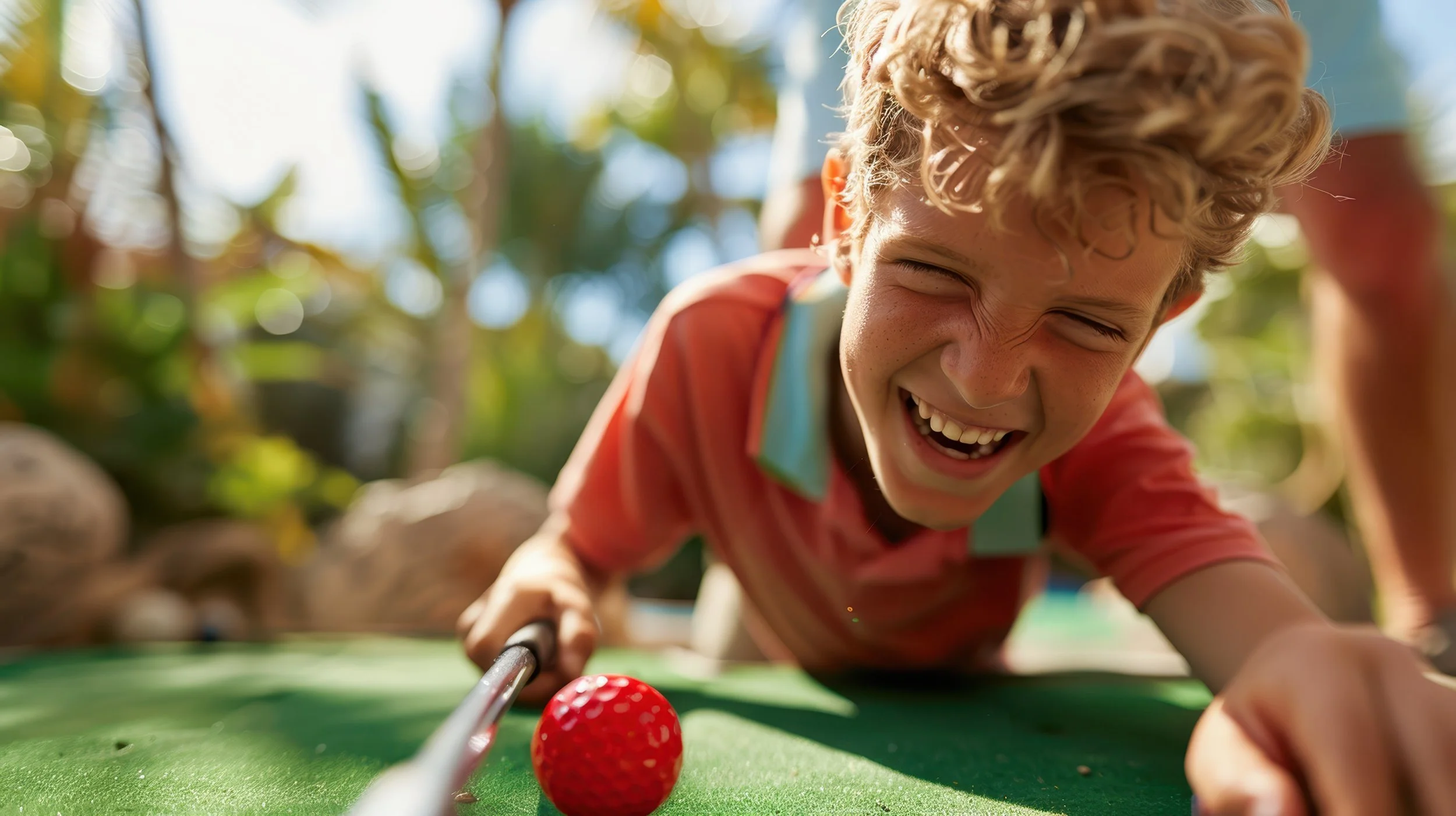 A young boy with curly blonde hair smiling and laughing as he plays mini-golf outdoors on a sunny day, leaning over a green golf course with a red golf ball in front of him.