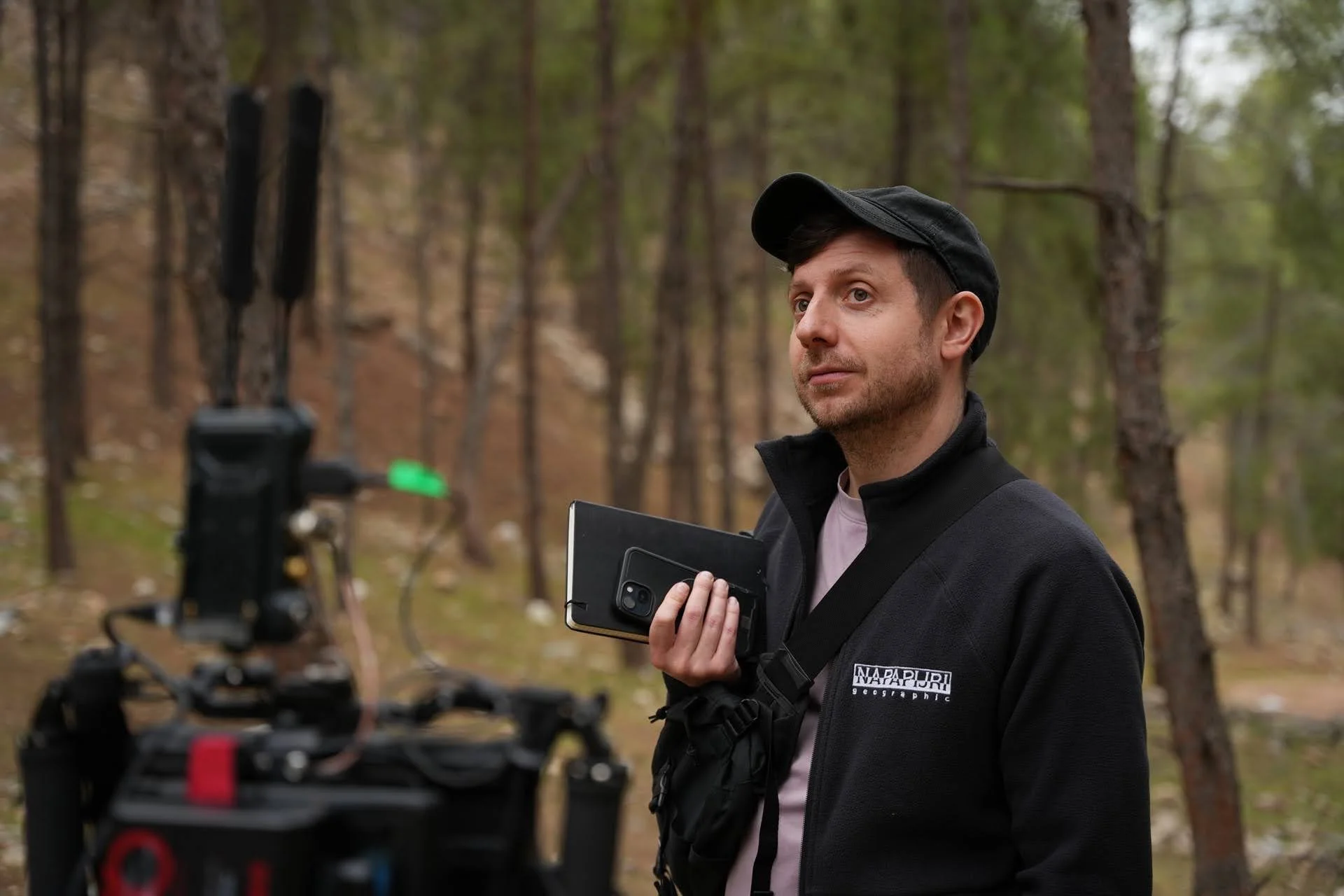 A man holding a camera and a tablet in a forest during the daytime, preparing for a filming or photography session.